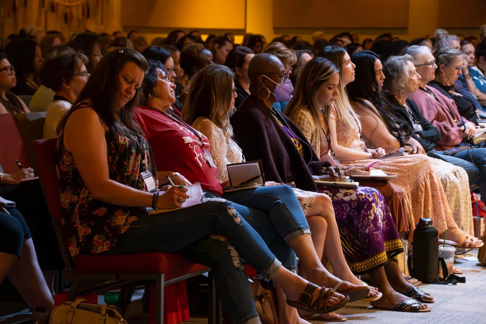 Audience of diverse women seated and listening attentively at a conference or seminar in a large room with wood-paneled walls.