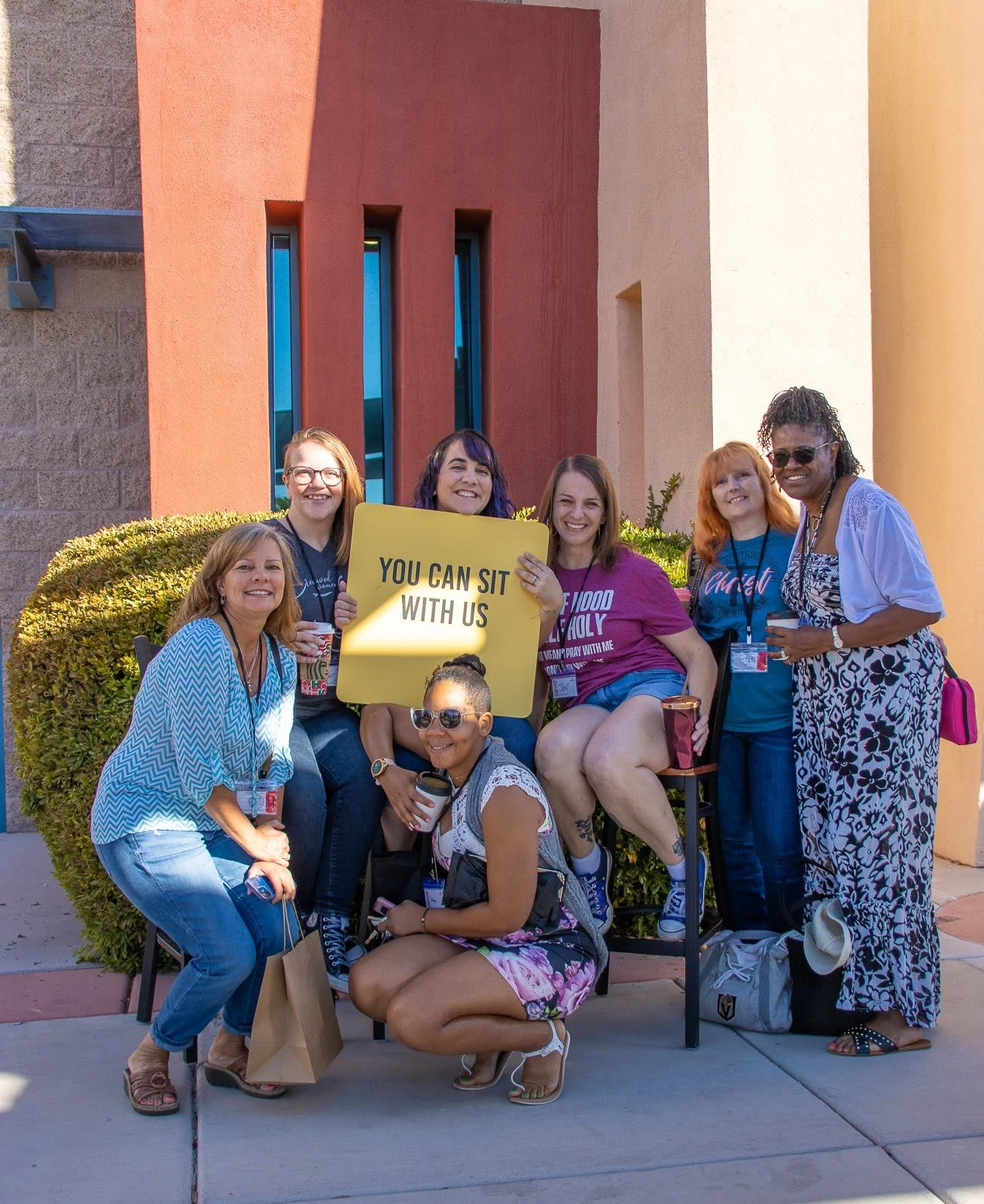 Group of diverse women and a girl gathered outdoors, holding a yellow sign that says "You Can Sit With Us," smiling, with colorful buildings in the background.