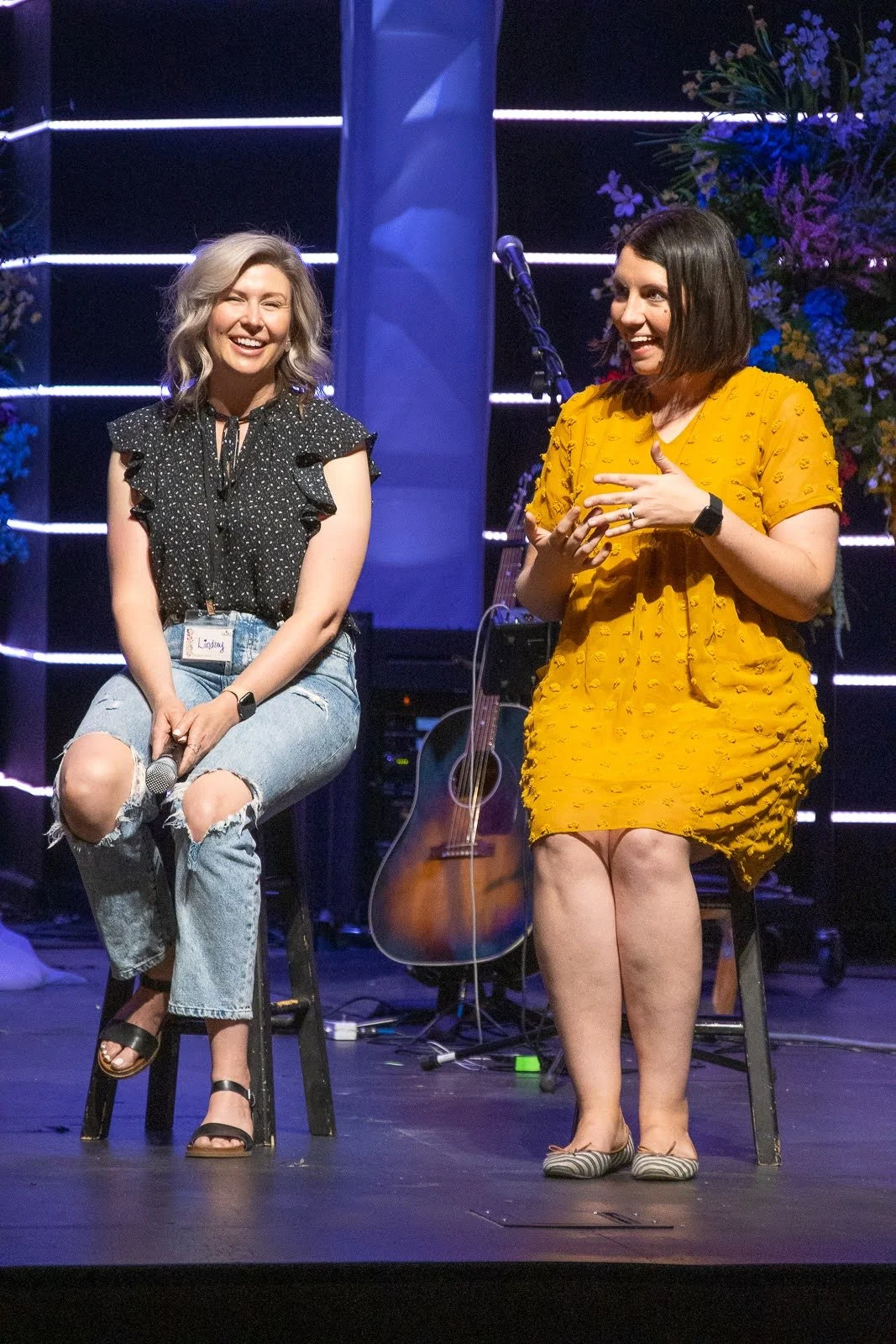 Two women on a stage, one sitting on a stool holding a microphone and smiling, the other standing and speaking, with a guitar and floral arrangements in the background.