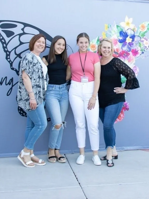 Four women standing side by side outdoors in front of a decorative butterfly and flower mural, smiling at the camera.