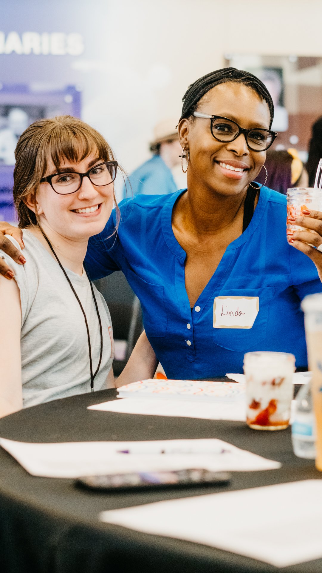 Two women smiling, one wearing glasses and a gray shirt, another wearing glasses and a blue shirt with a name tag that says "Linda," at a table with food containers and papers, in a social or professional setting.