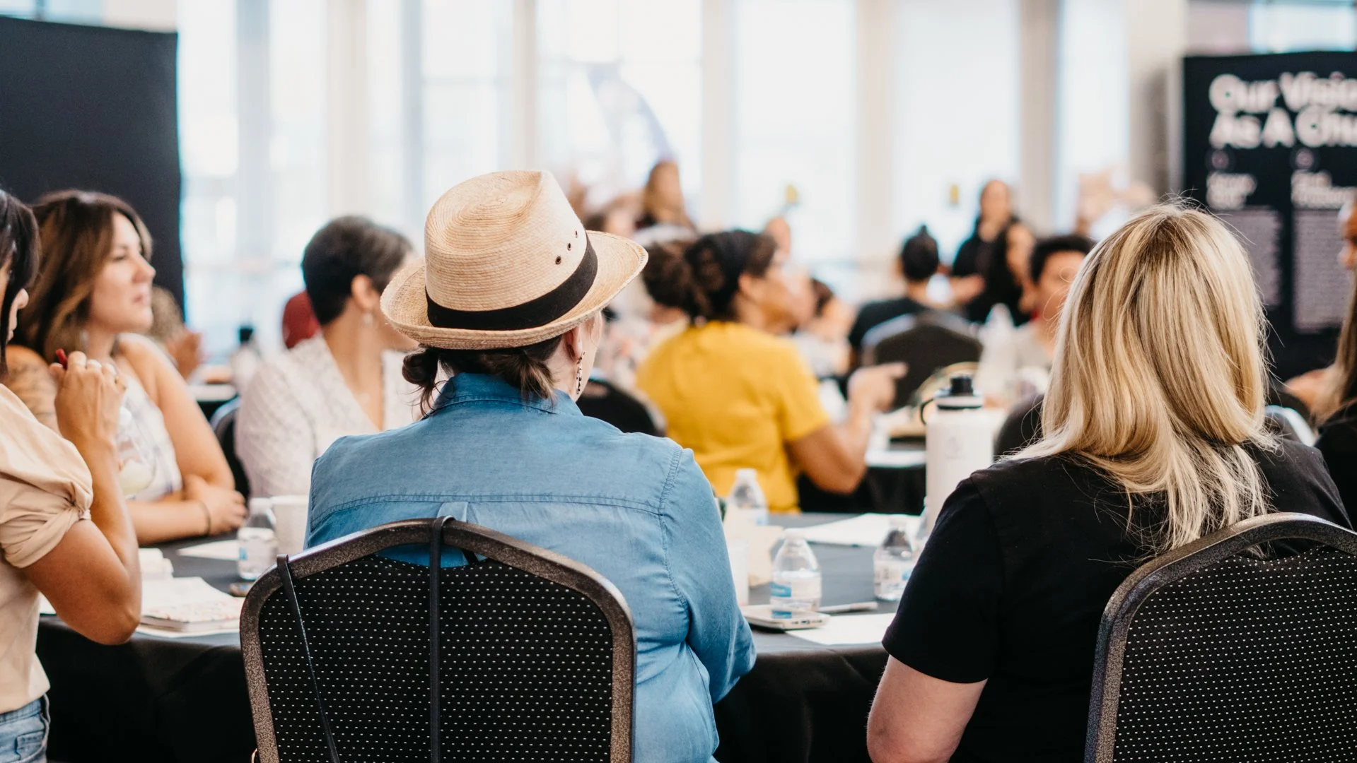 A group of people attending a conference or seminar in a large, well-lit room with floor-to-ceiling windows. The audience features women and men seated at tables, listening to a speaker at the front. Some are taking notes, drinking water, and looking attentively.
