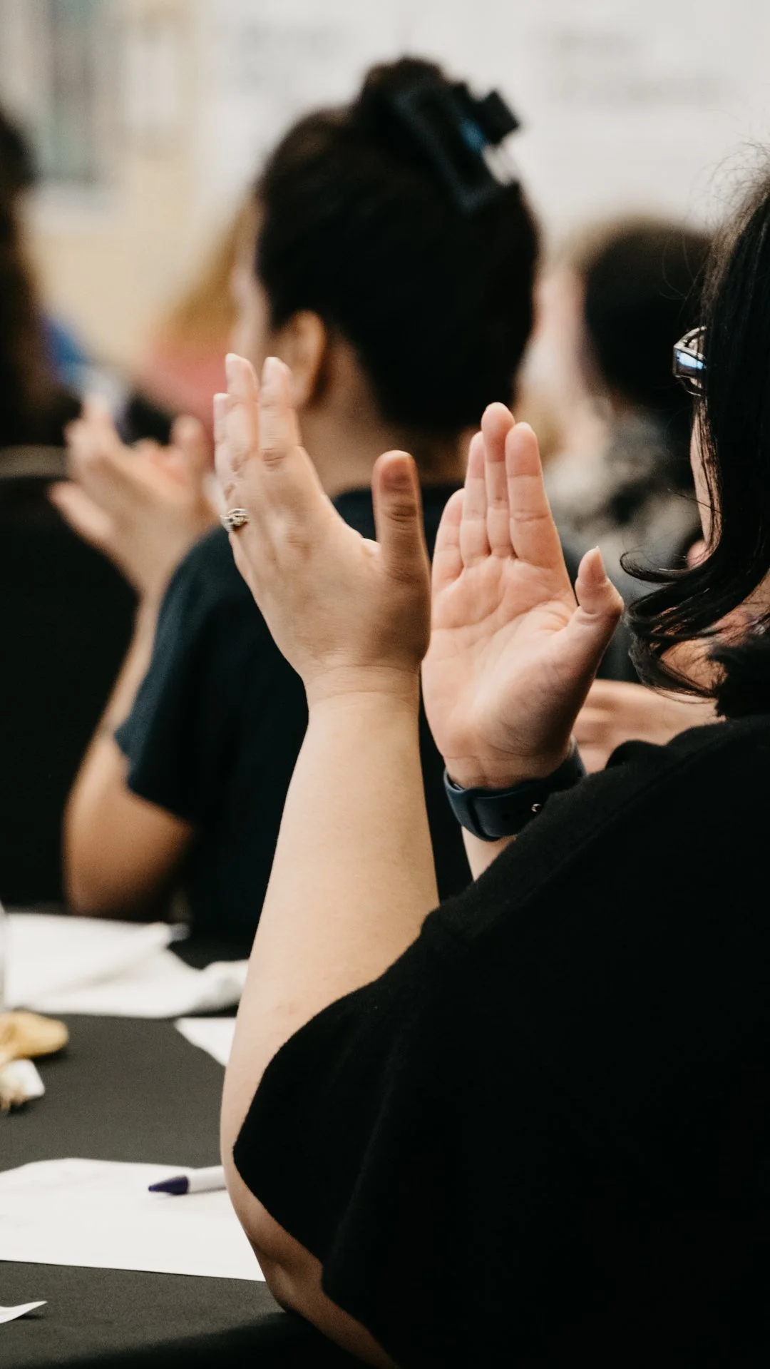 Close-up of a person's hands raised, clapping during a gathering or event, with other attendees in the blurred background.