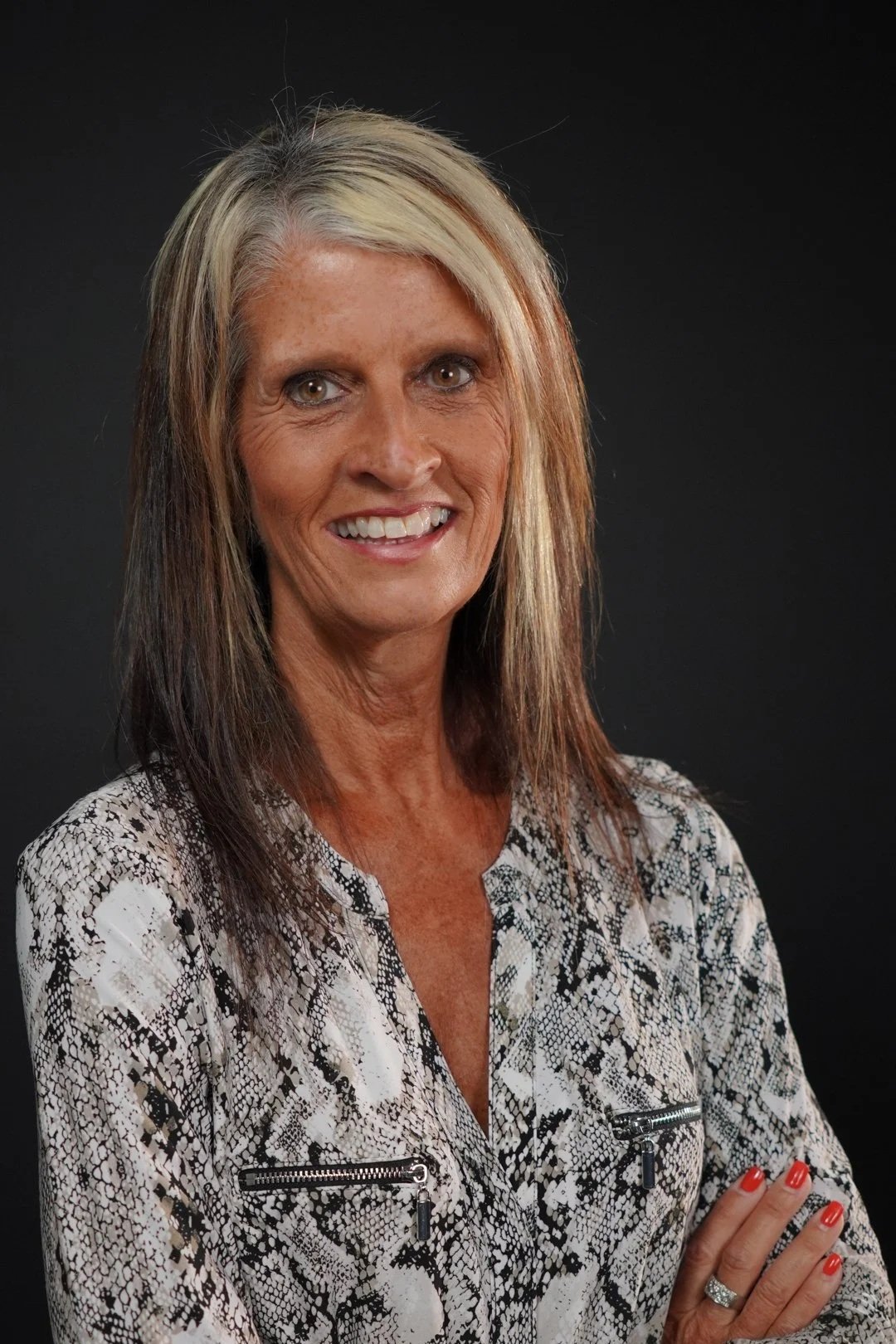A smiling middle-aged woman with shoulder-length blonde and brown hair, wearing a patterned blouse with zippers on the chest, standing against a black background.