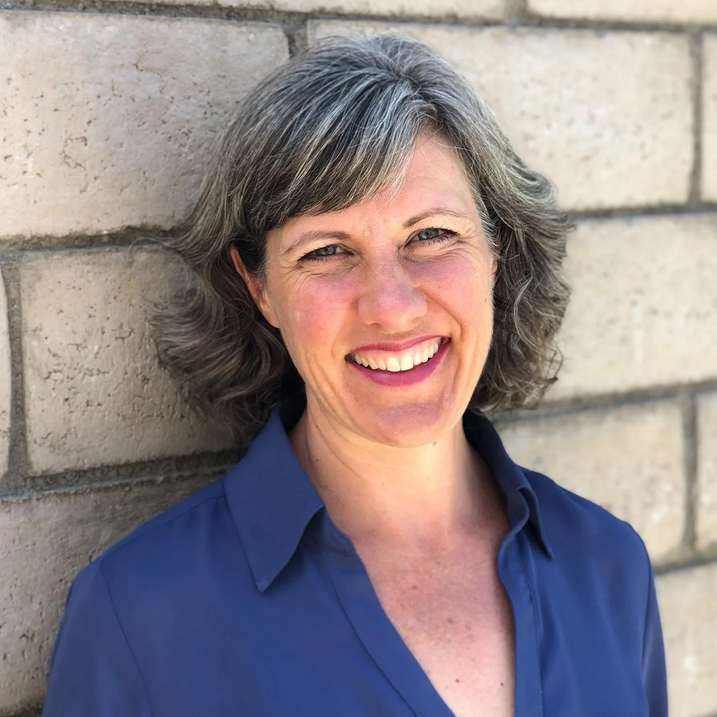 A middle-aged woman with short gray hair, blue eyes, and fair skin, smiling and wearing a blue shirt, standing against a beige brick wall.