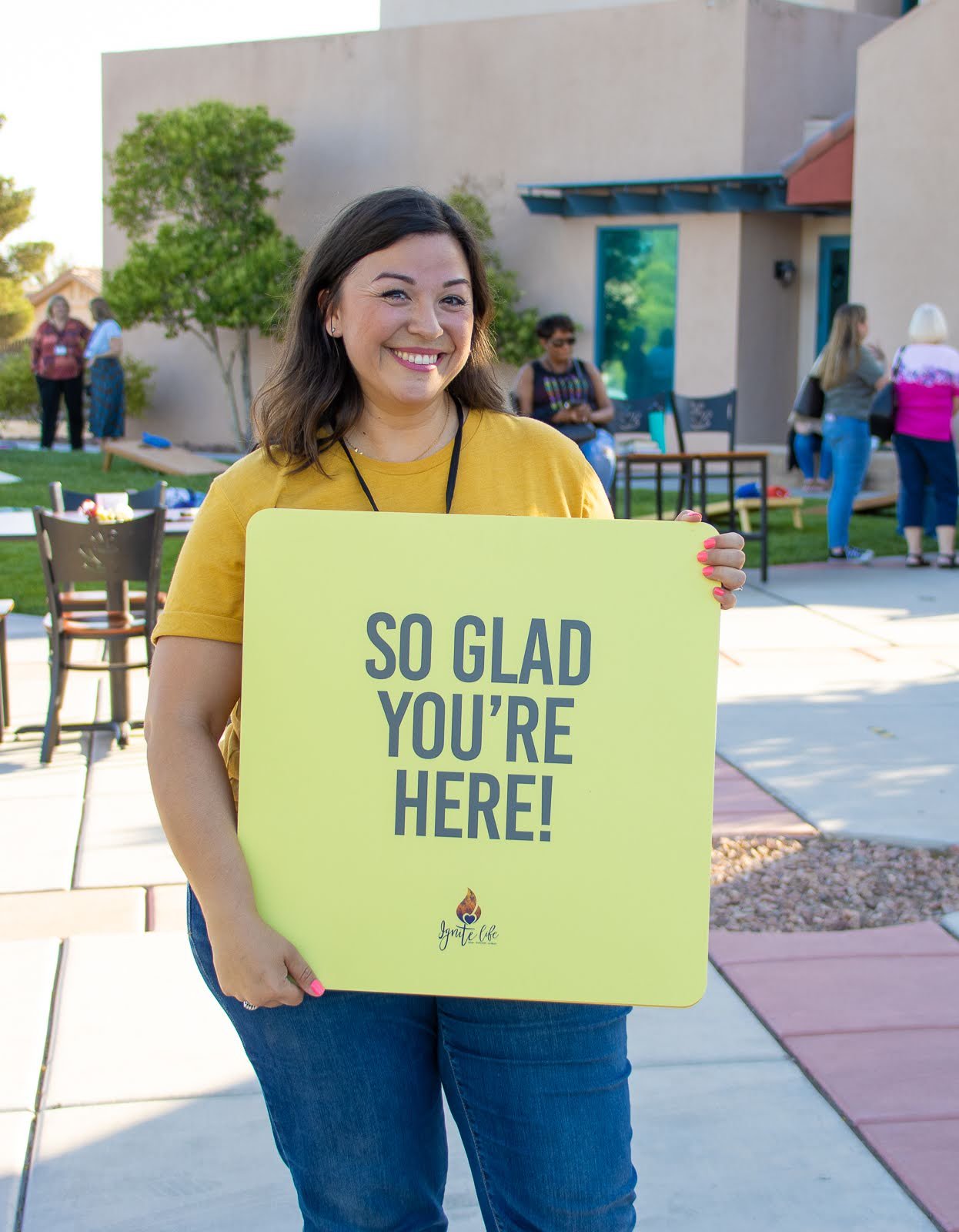 A smiling woman in a yellow shirt holding a yellow sign that reads "So glad you're here!" at an outdoor gathering or event with several people in the background.