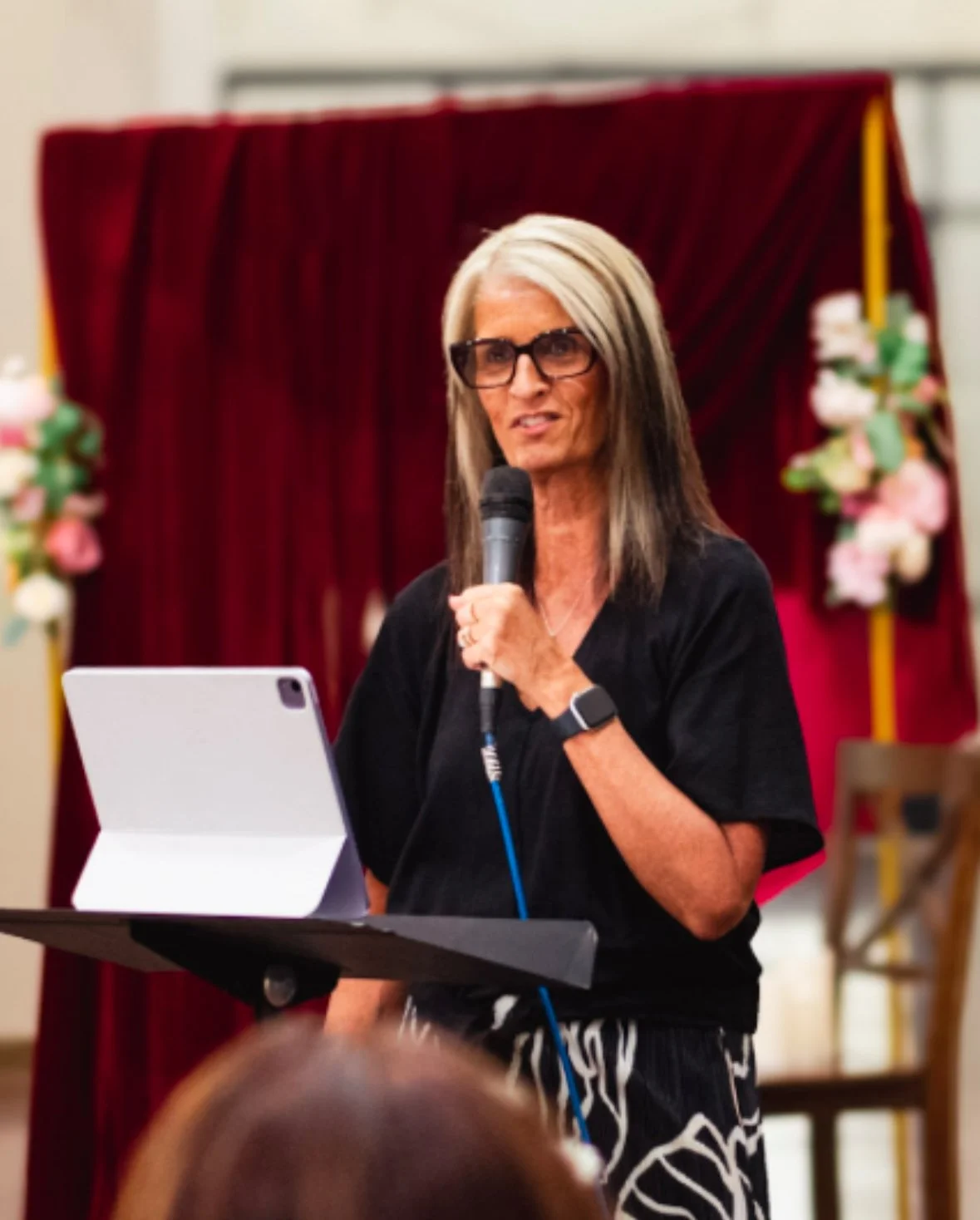 A woman with blonde and brown hair, wearing glasses and a black shirt, speaking into a microphone. She is standing near a small table with a tablet, with a red curtain and floral decorations in the background.