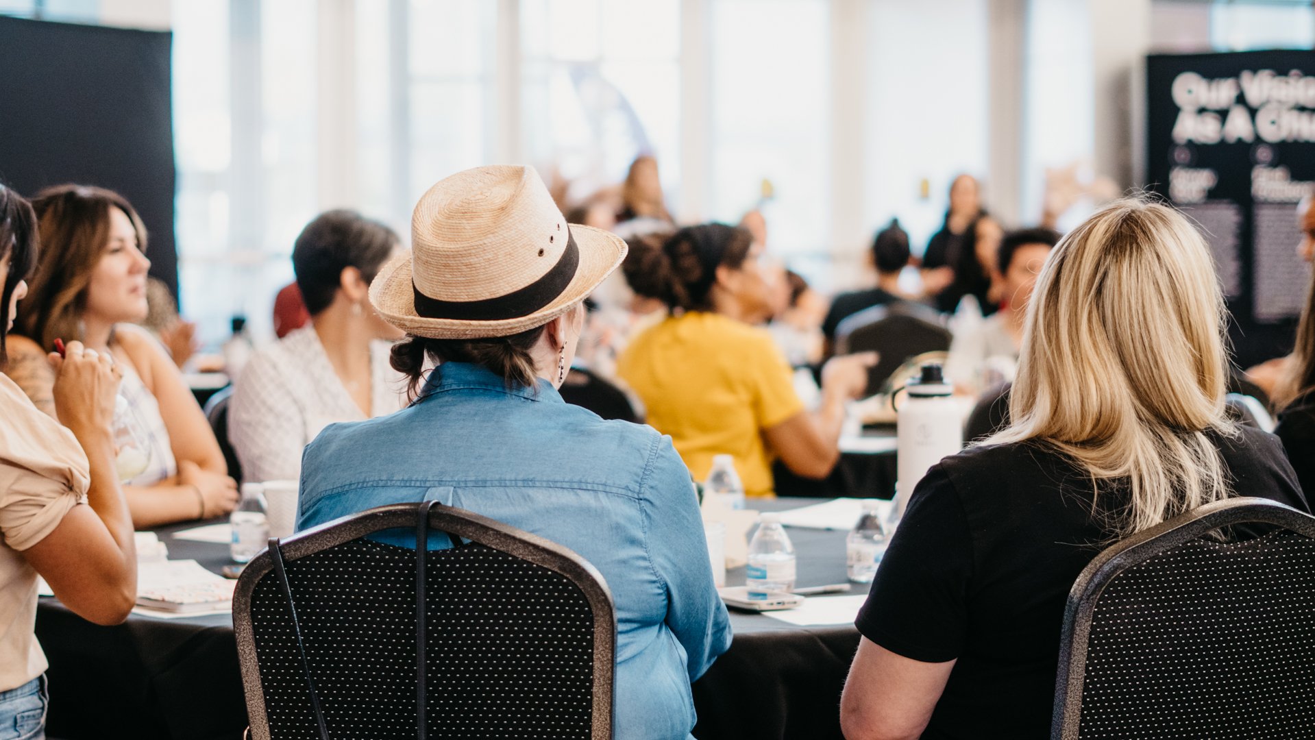 A diverse group of people attending a conference or seminar in a large, well-lit room with large windows, sitting around tables with bottled water and note-taking supplies.