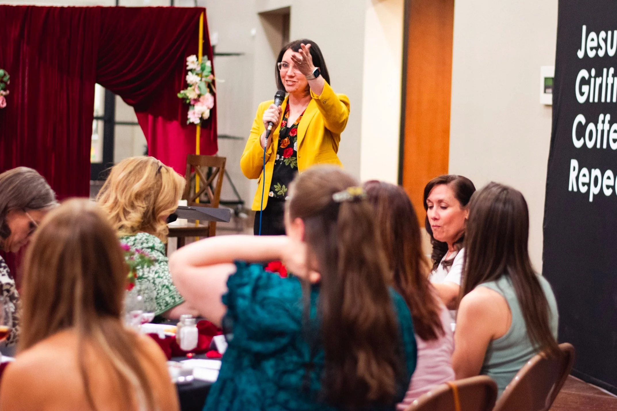 A woman in a yellow blazer stands at the front of the room, speaking into a microphone and gesturing with her hand, while an audience listens. The setting appears to be a conference or seminar with a decorative backdrop of flowers and a partially visible blackboard with white text.