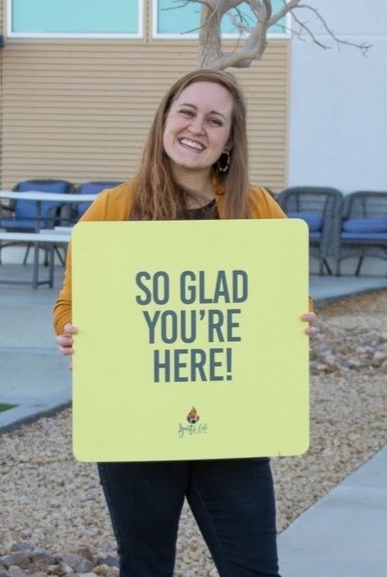 A woman smiling and holding a yellow sign that says, "SO GLAD YOU'RE HERE!" outside in front of a building with windows and benches.