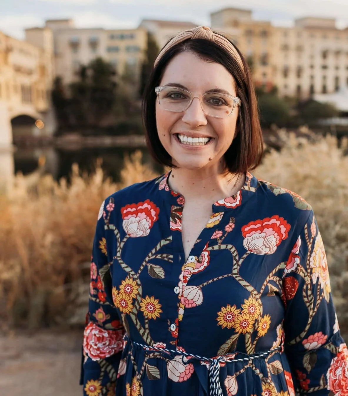 A smiling woman with short dark hair, wearing glasses and a headband, in front of a cityscape with buildings and a river, during golden hour.