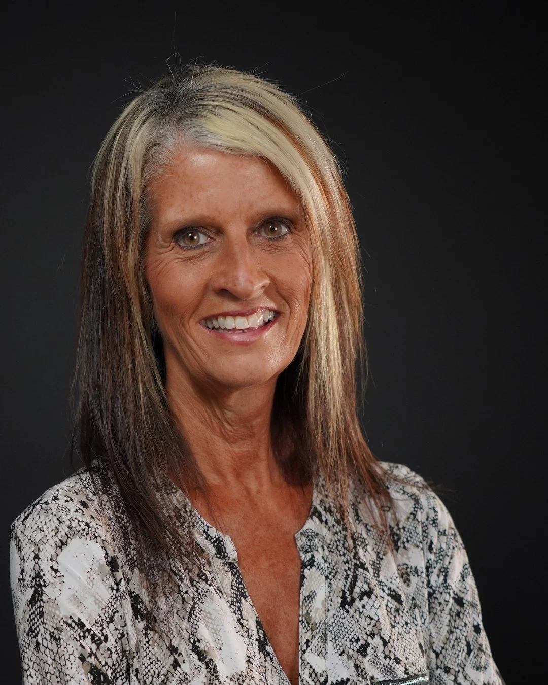 Portrait of a middle-aged woman with long, light brown and blonde hair, smiling, wearing a patterned blouse, against a dark background.