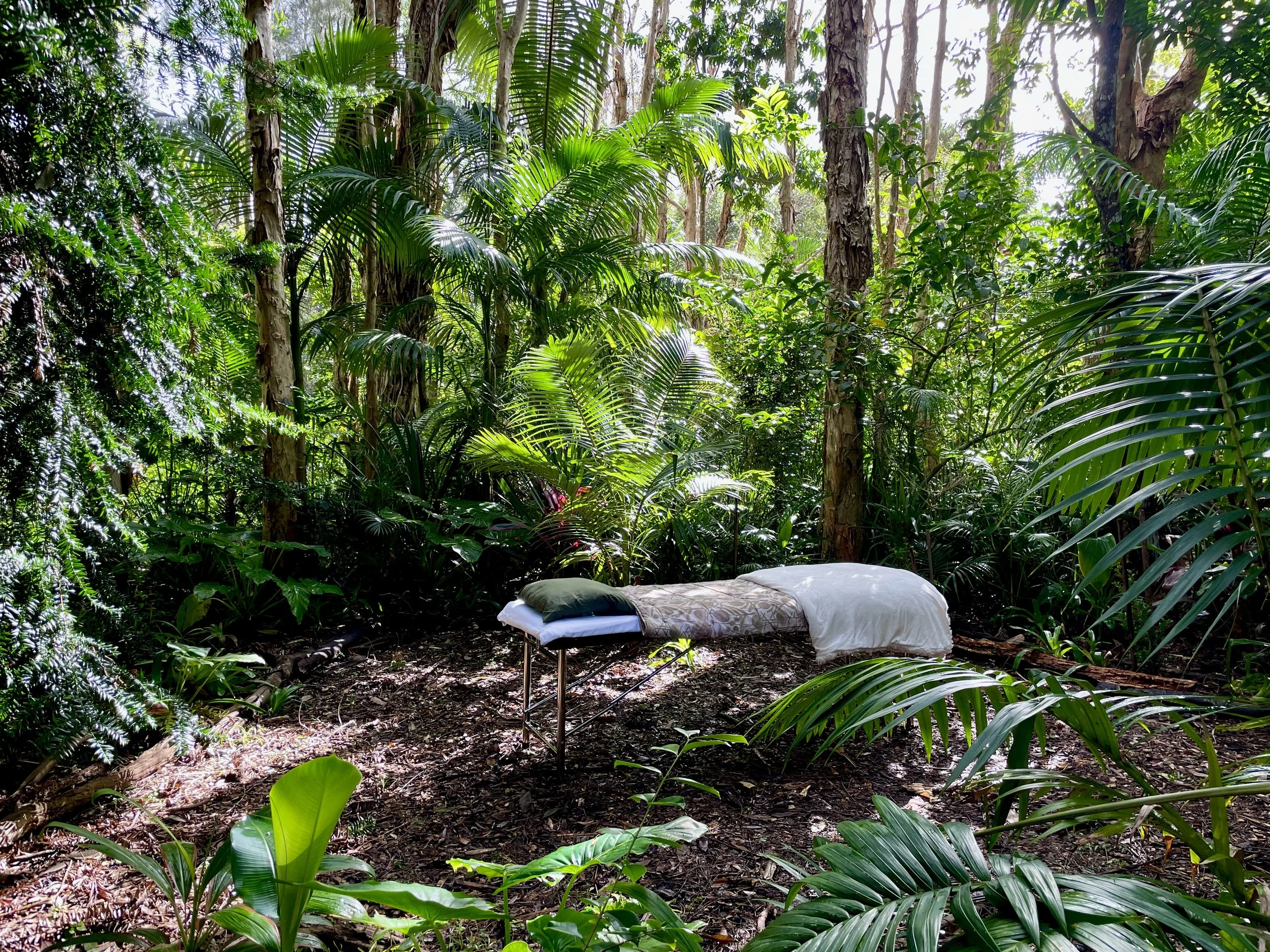 A portable massage table with white pillow and blanket set up in a lush, green tropical forest, surrounded by tall trees and dense foliage.