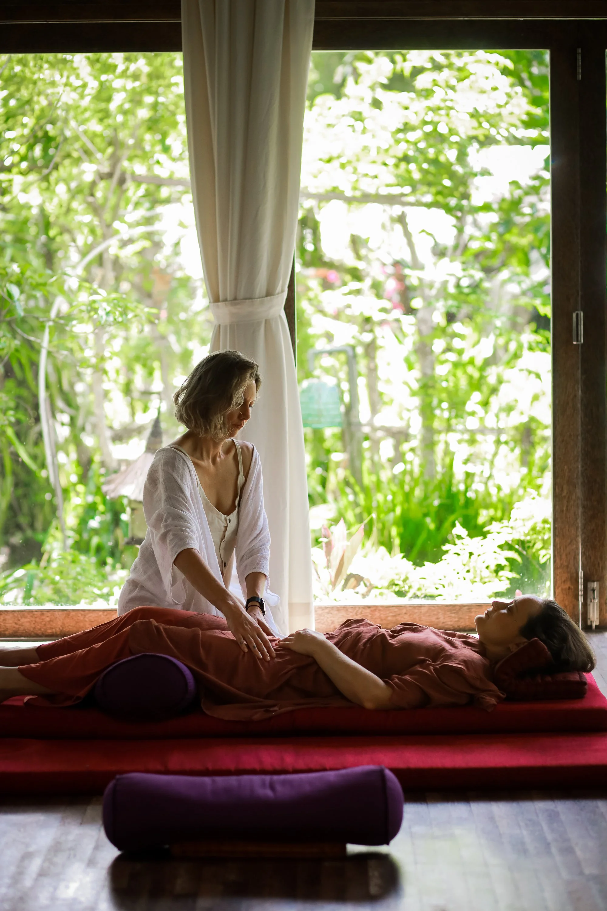 A woman giving a massage to a person lying on a bed in a room with large windows showcasing greenery. The woman stands by the bed, gently pressing on the person's chest, while the person reclines with their eyes closed.