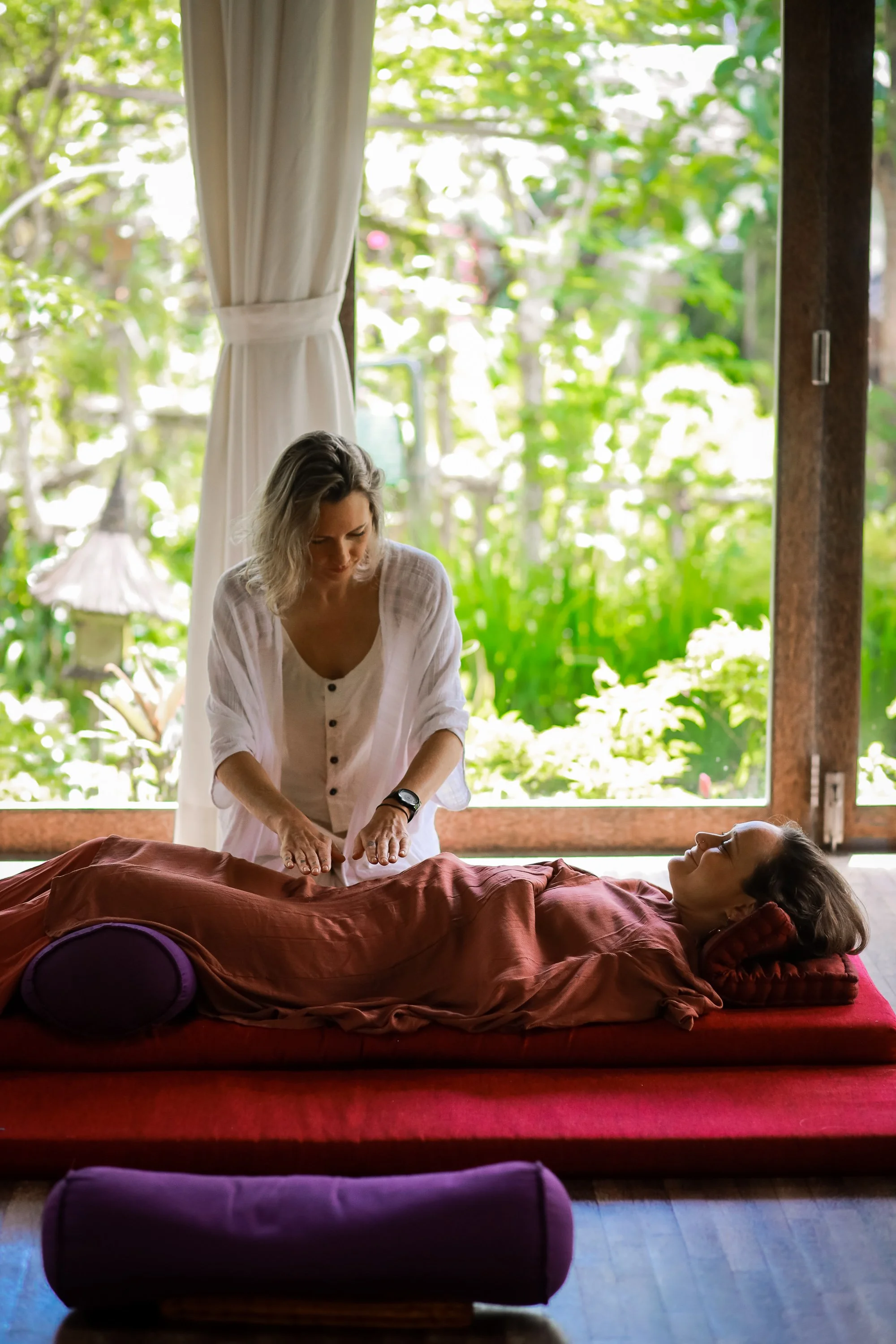 A woman receiving a massage from a massage therapist in a room with large windows and lush greenery outside.