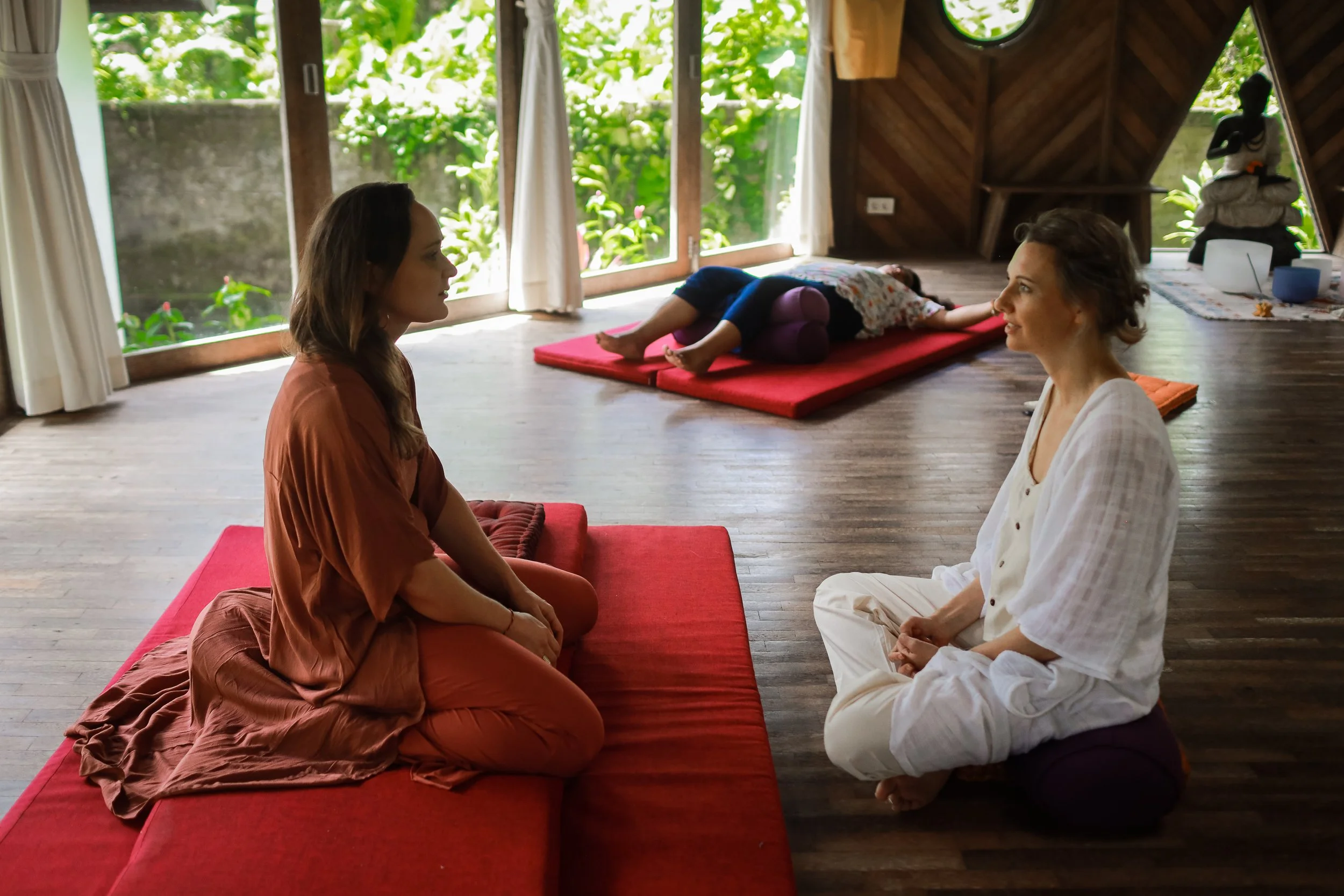 Two women sitting face-to-face on a red cushion with their legs crossed, engaging in a meditation or conversation inside a wooden room with large windows revealing green foliage outside. A person is lying on a mat in the background, and there is a statue and some meditation supplies in the corner.
