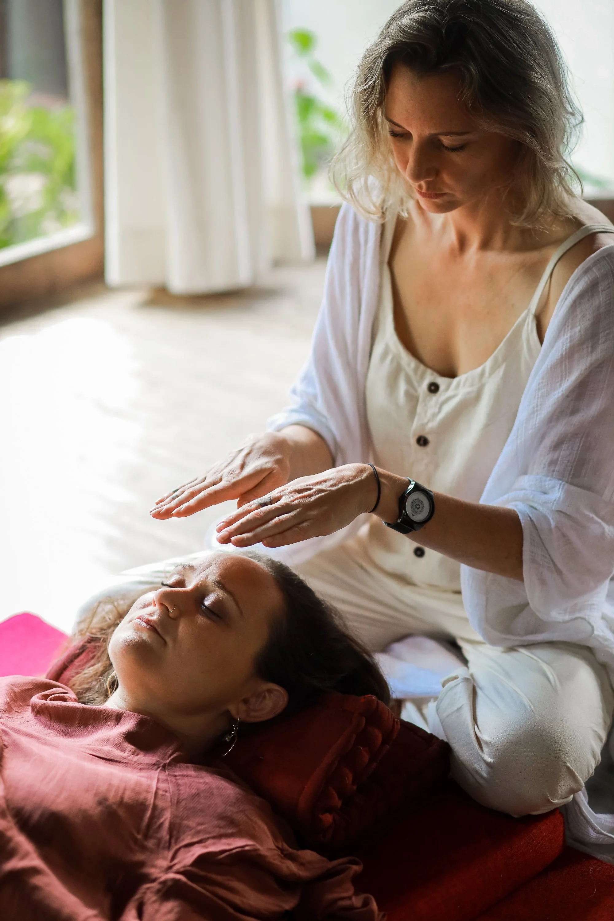 A woman gives a massage or healing treatment to a young woman lying on a bed with her eyes closed, in a bright room with natural light and green plants outside the window.