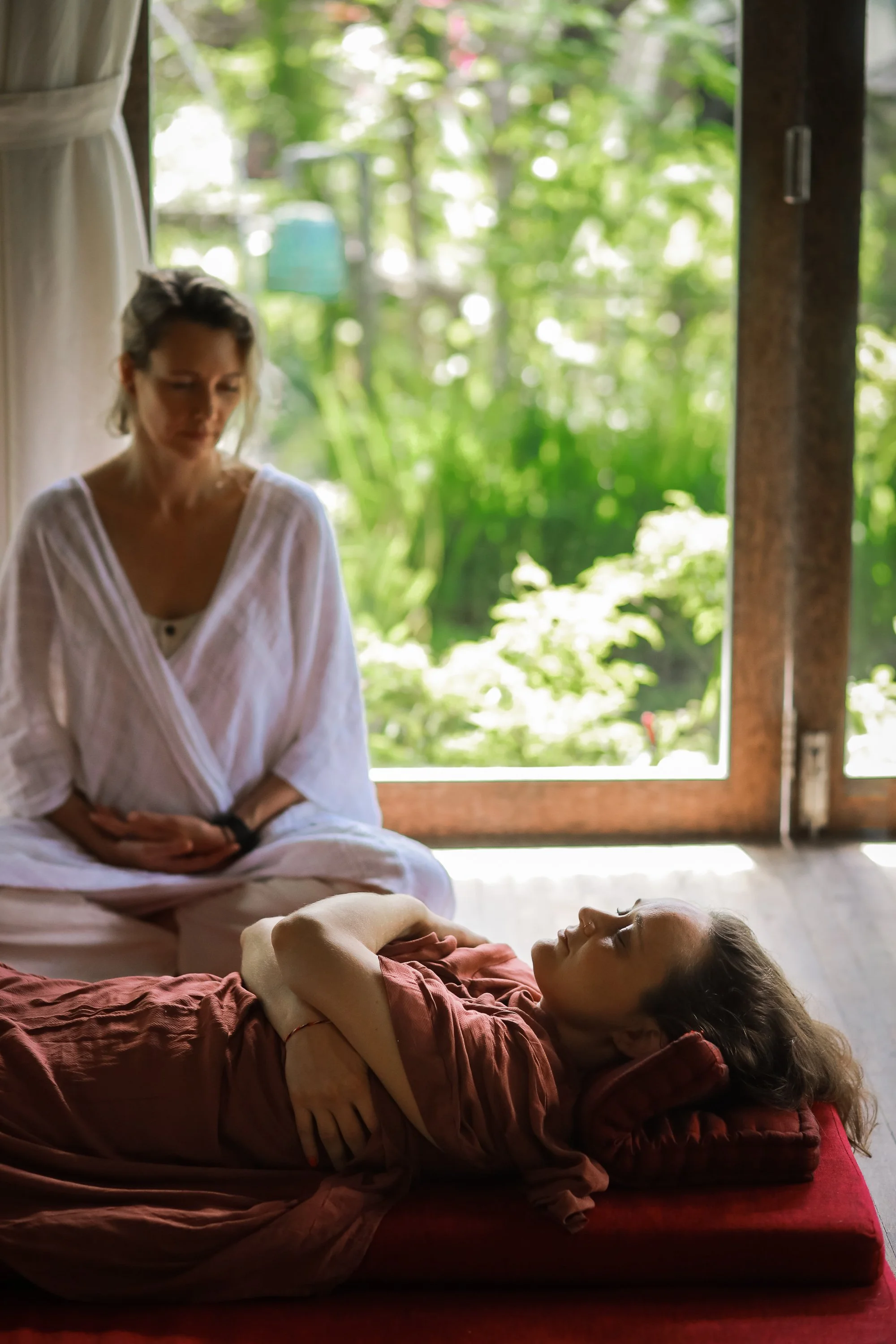 A woman in white attire and a young girl in a brown dress are practicing yoga or meditation together, indoors near a large window showing green foliage outside. The girl is lying on her side on a cushion, with her eyes closed and arms crossed over her chest. The woman is sitting cross-legged, watching the girl, with her hands resting in her lap.