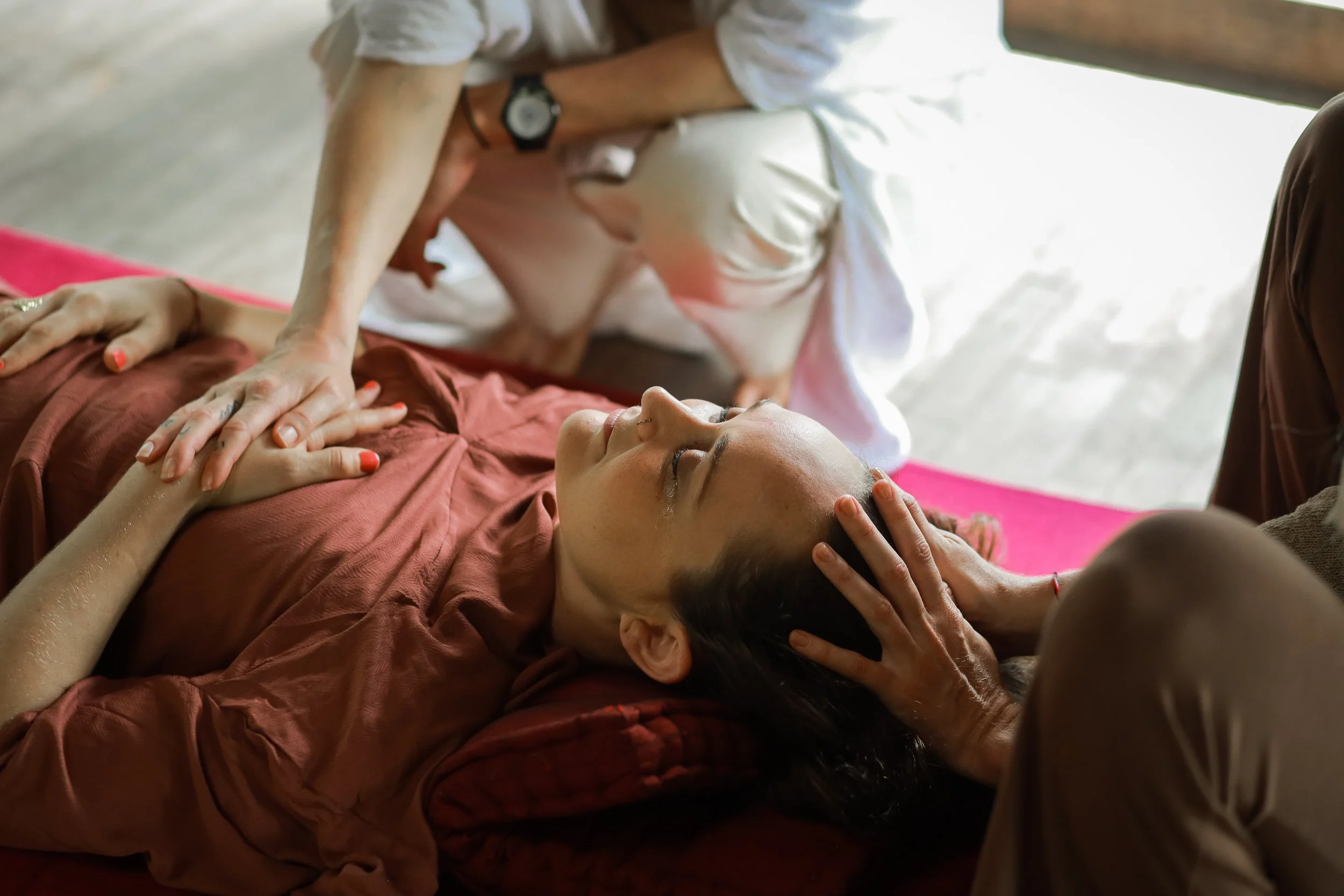 A person lies on their back with eyes closed, while two others lean over them giving a massage on a pink yoga mat. The scene suggests a relaxing or therapeutic environment.