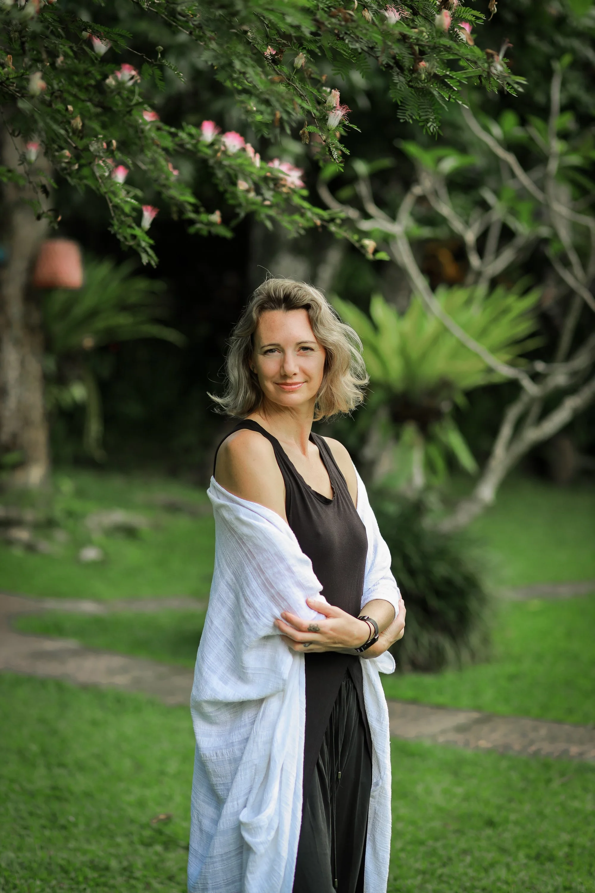 A woman with blonde hair and a slight smile stands outdoors in a green park or garden, wearing a black dress and a white cardigan. She is crossing her arms, with a lush background of trees and plants.