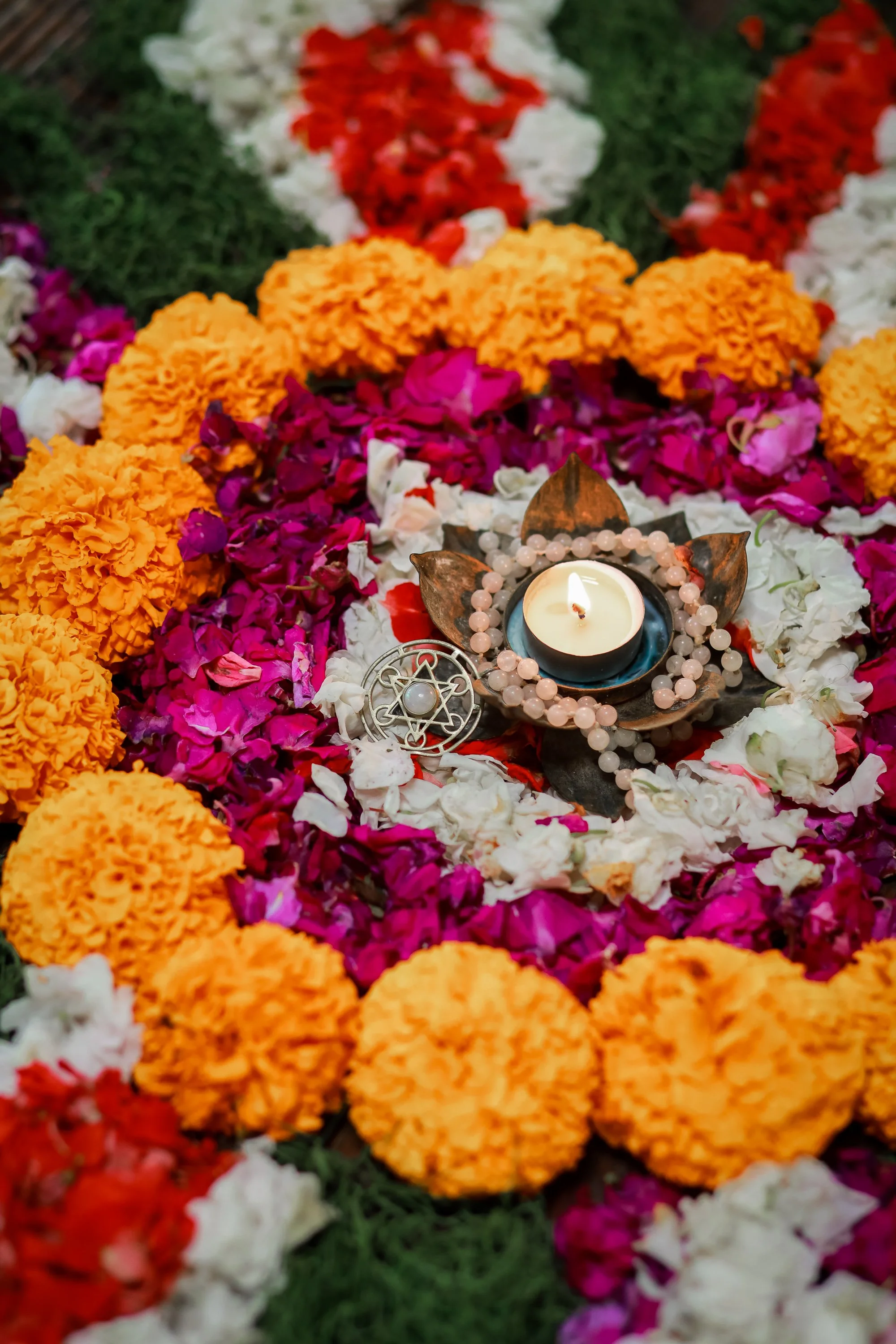 A memorial surrounded by colorful flower petals, with a lit candle, beads, and metallic ornaments.
