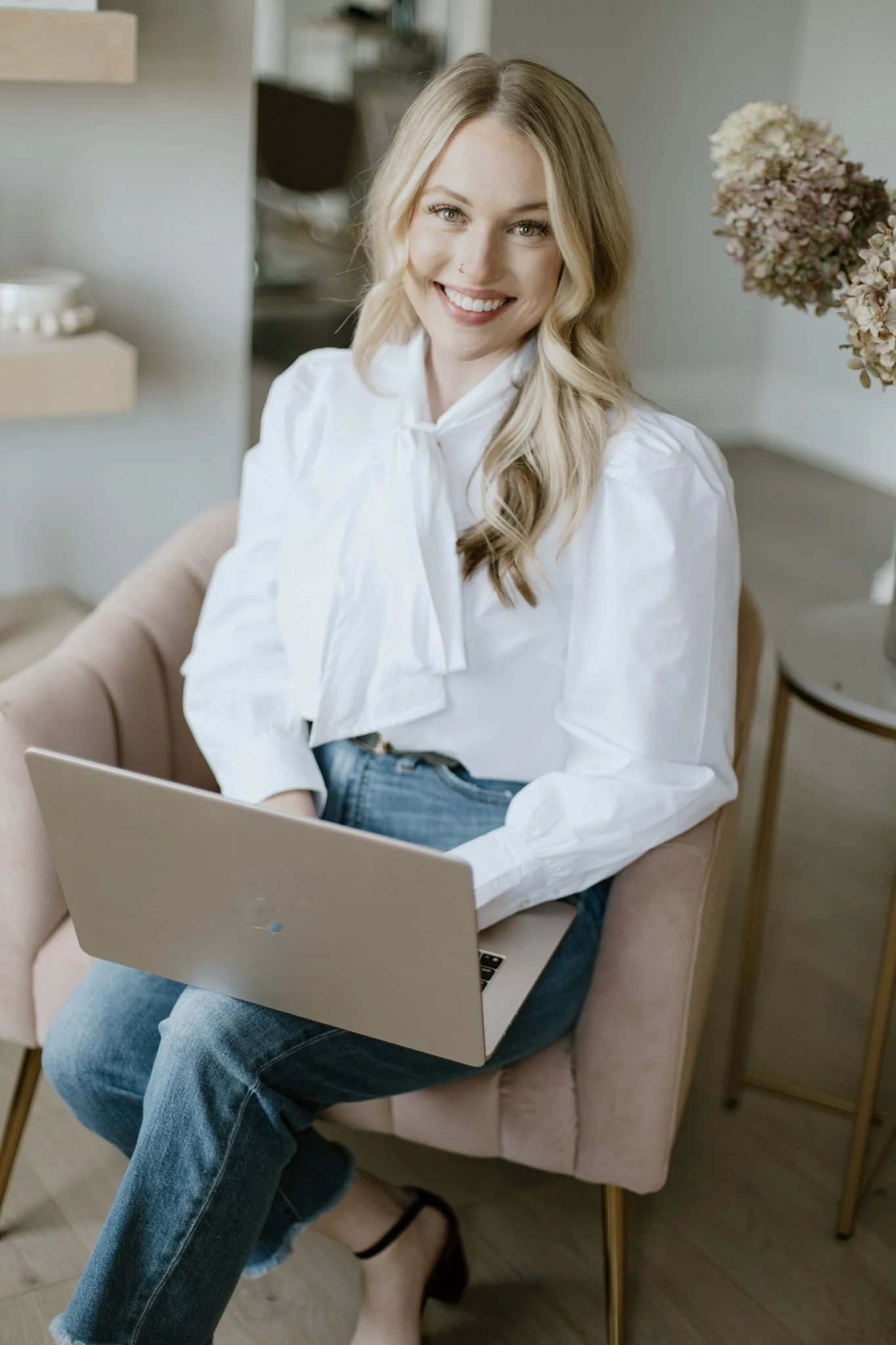 A woman sitting on a pink armchair using a laptop, with a laptop on her lap, wearing a white blouse and blue jeans, in a cozy indoor setting with flowers and shelves in the background.