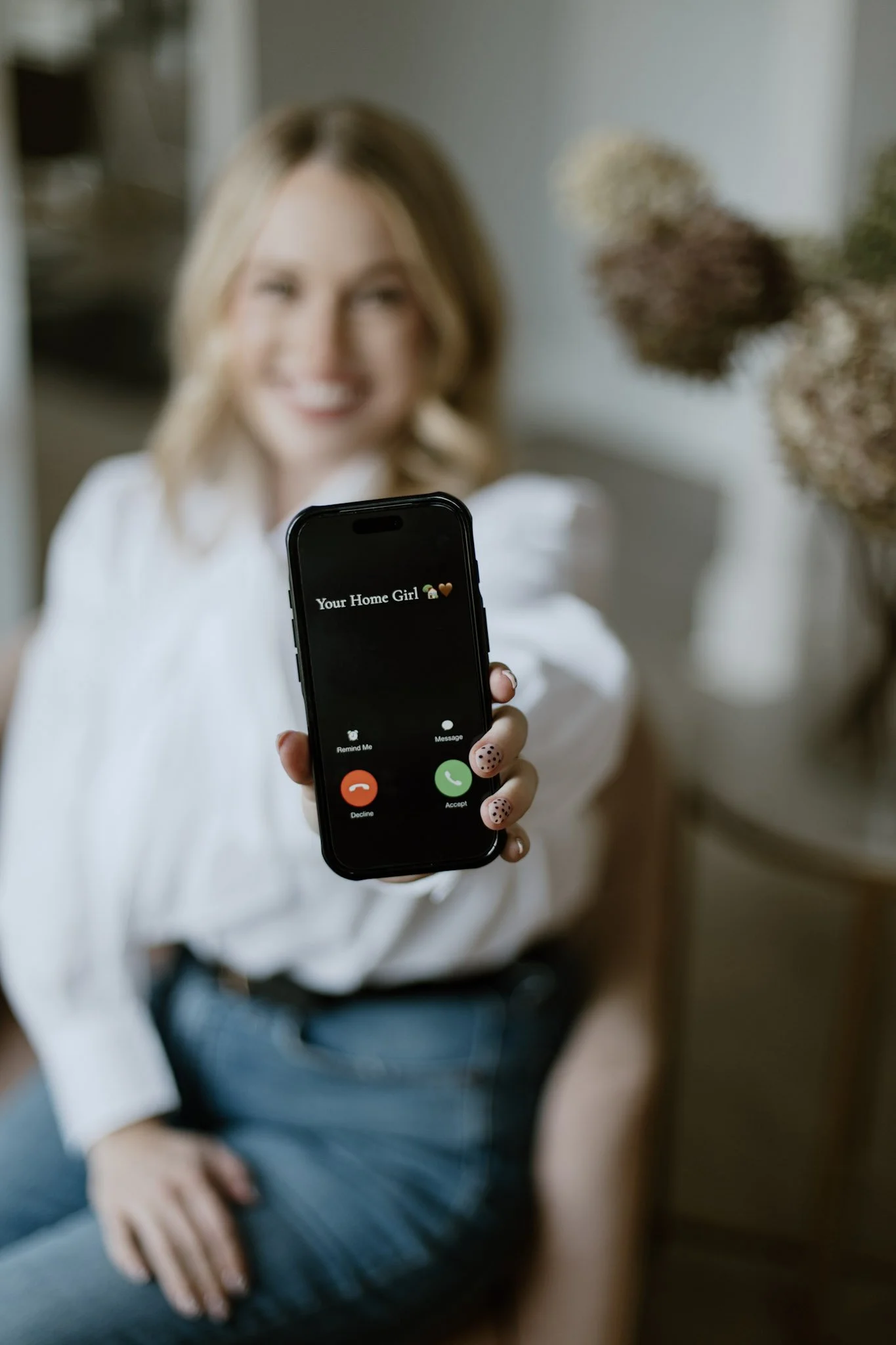 A woman sitting on a couch holding a smartphone displaying an incoming call from 'Your Home Girl' with a lock icon and a heart emoji, smiling in the background.