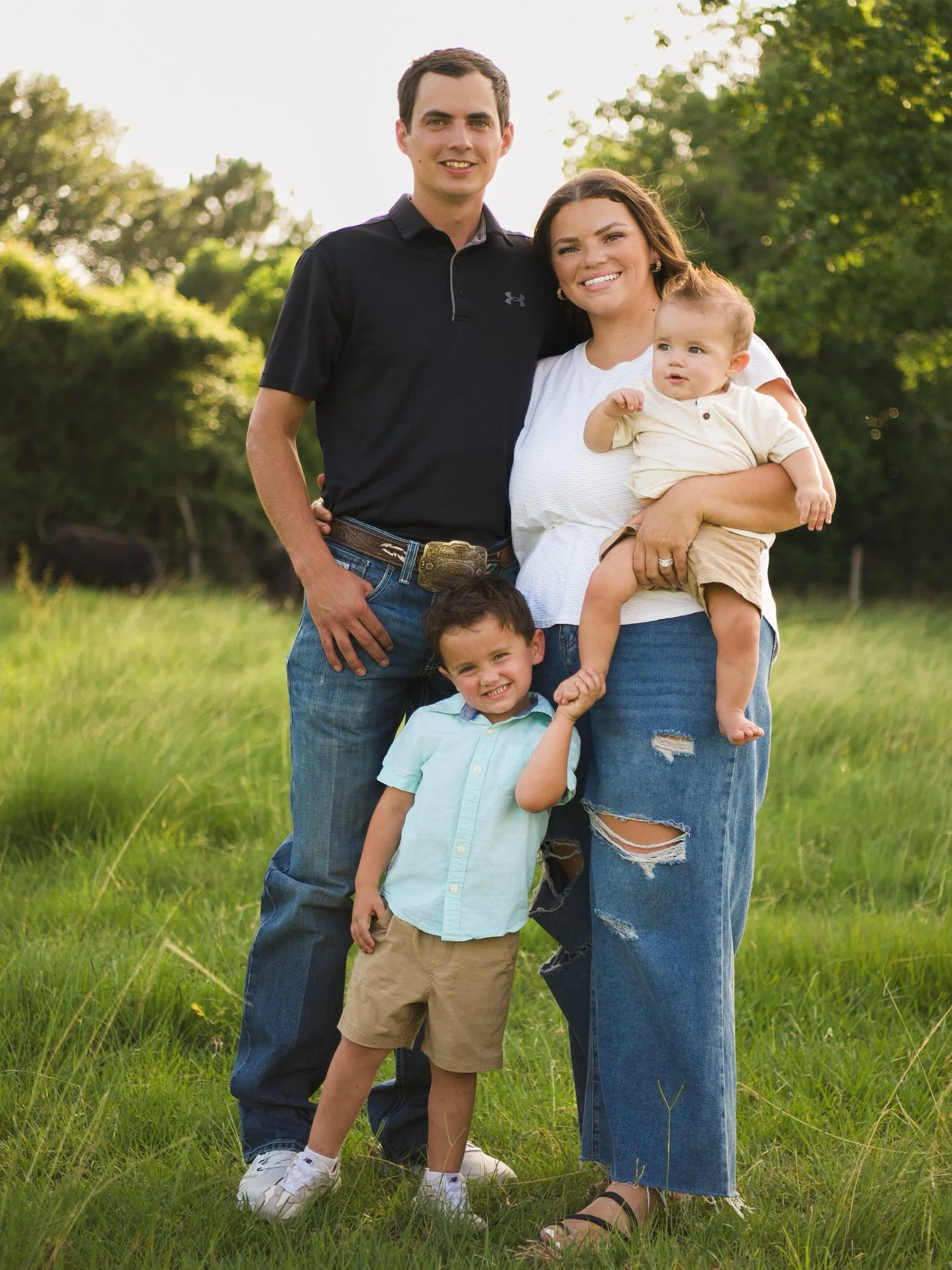 A family of five standing on grass in a park during daylight, with trees in the background. The father wears a black polo shirt and jeans, the mother wears a white top and ripped jeans, a young boy in a light blue shirt and khaki shorts is in front, and a baby with a cream shirt is being held by the mother. Another child, partially visible, is at the bottom.