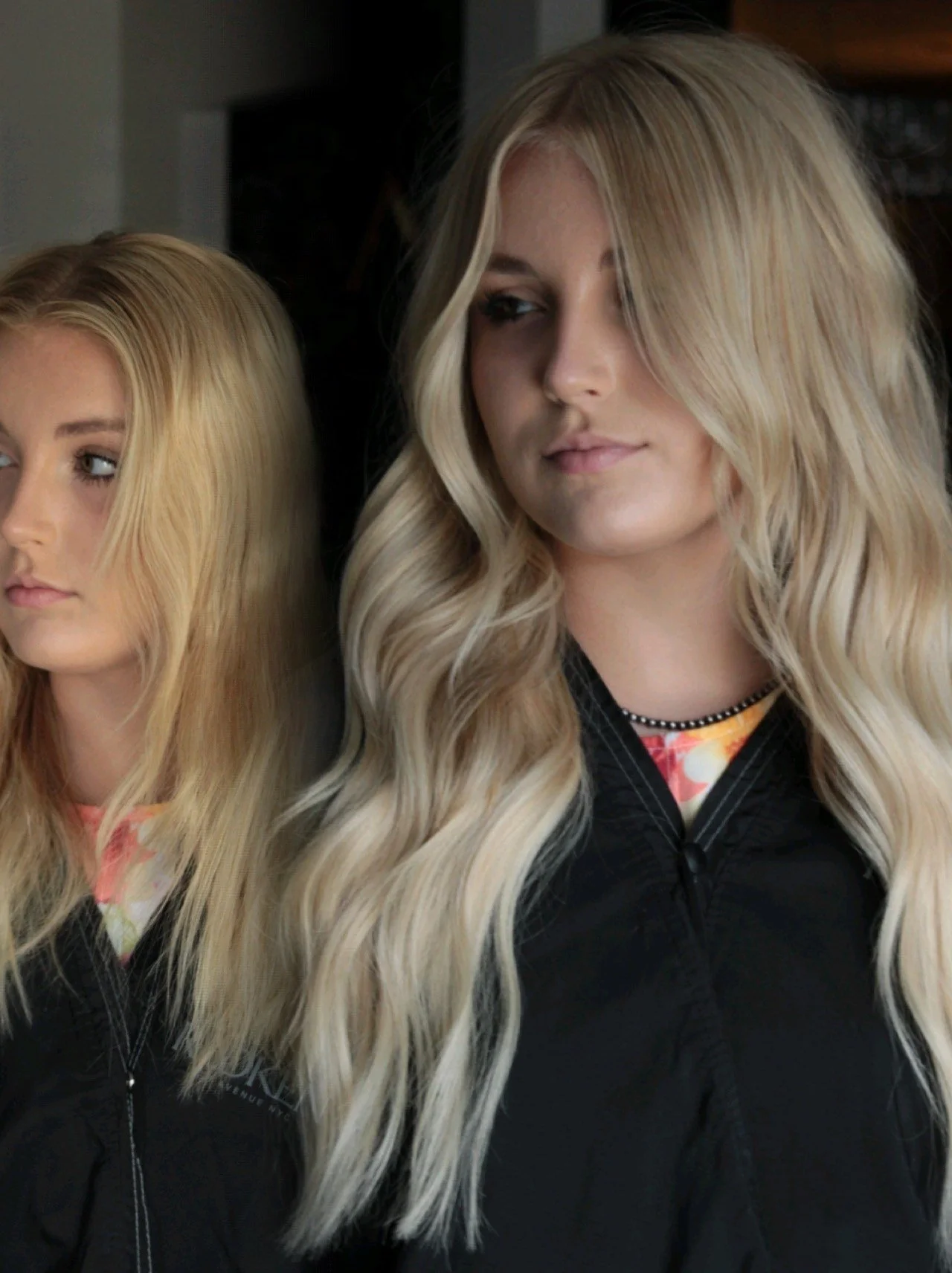 Two women with long blonde hair standing next to each other, wearing black jackets.