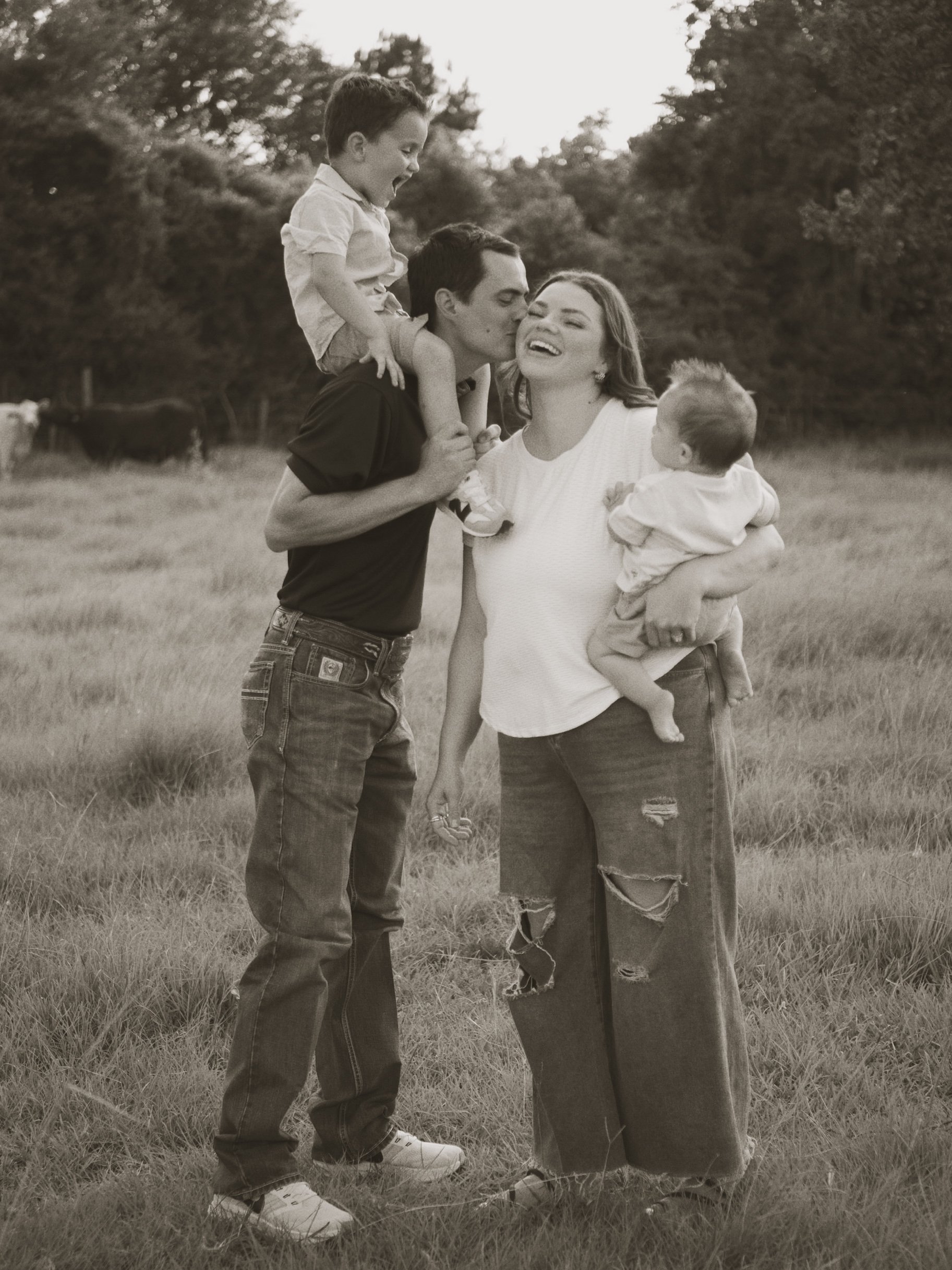 A family of four outdoors on a grassy field with trees in the background. The father is carrying his older son on his shoulders, who is laughing. The mother, holding their younger child, is smiling. The scene appears joyful and playful.