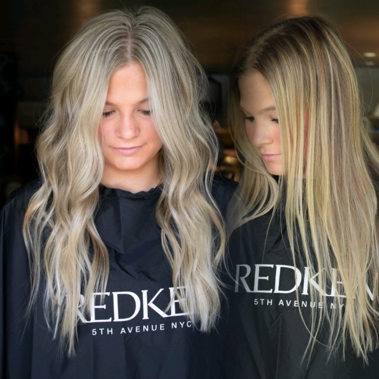 Two women with long blonde hair, wearing black salon capes with the text "REDKEN 5TH AVENUE NYC", standing close together with heads slightly bowed and smiling gently in a salon.