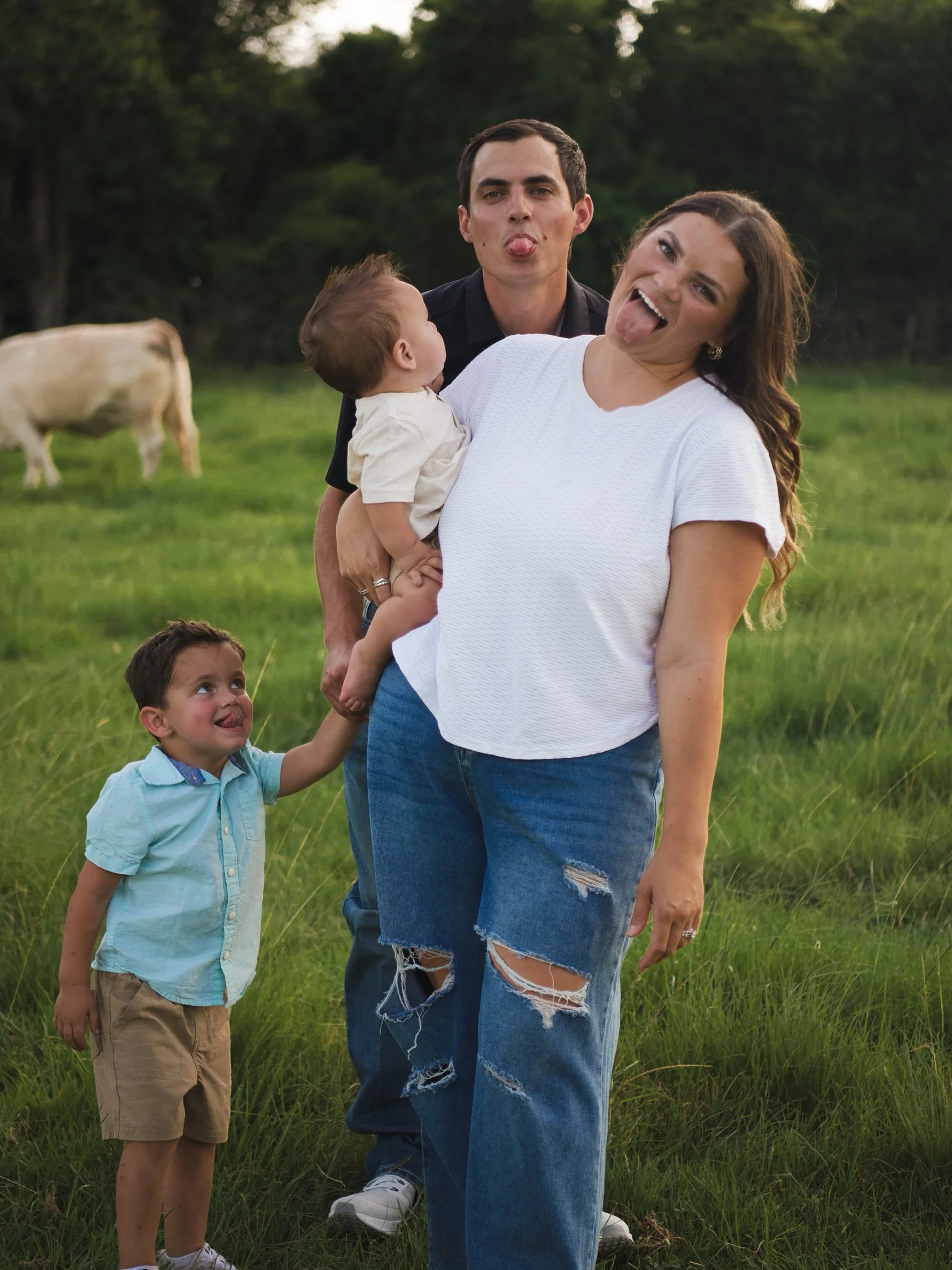 A family of four outdoors in a grassy field, making funny faces. The mother is wearing a white shirt and ripped jeans, the father is behind her with his tongue out, the son is holding her hand and looking up, also with his tongue out, and the baby is held by the father, facing the mother.
