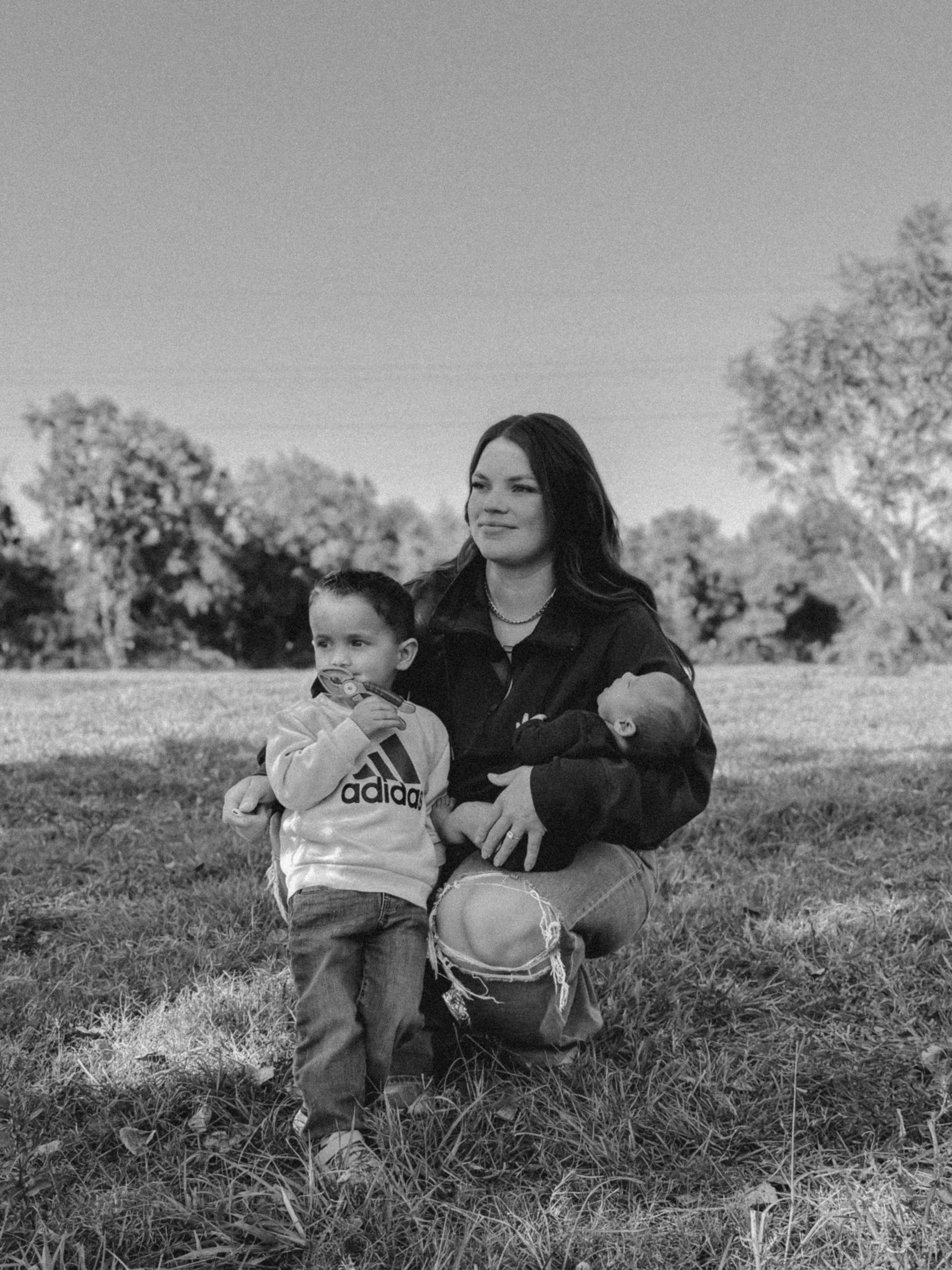 A woman sitting on the grass outdoors with two young children, one standing and one lying on her lap, with trees in the background on a clear day.