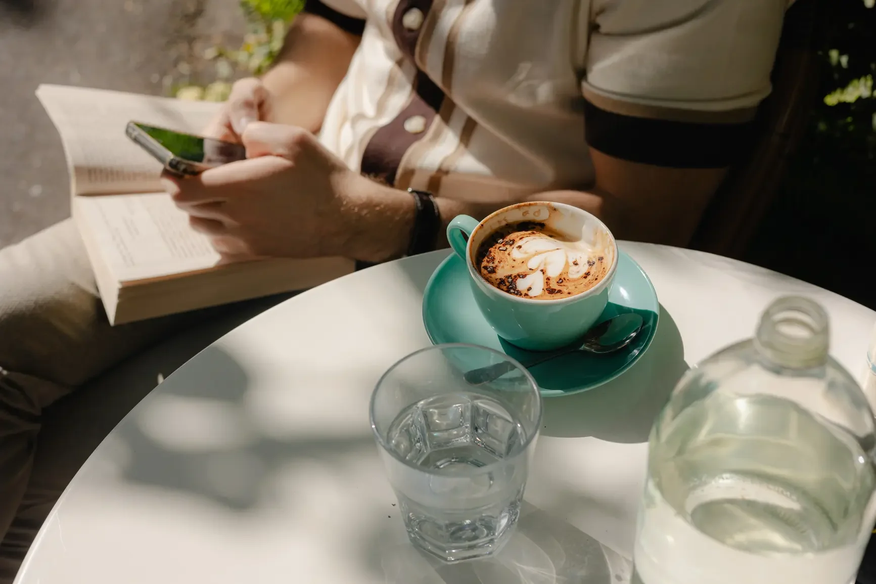 A person sitting at a white table, reading a book and holding a smartphone, with a cup of coffee topped with whipped cream and chocolate, a glass of water, and a water bottle on the table.