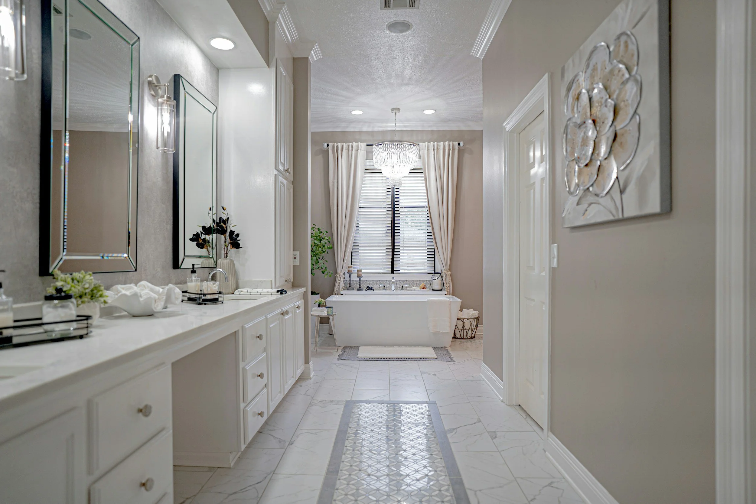 Bright bathroom with white vanity, mirrors, and decorative light fixtures. A bathtub with curtains, a chandelier, and a window with blinds are visible in the background. The floor has marble tiles with a patterned border.