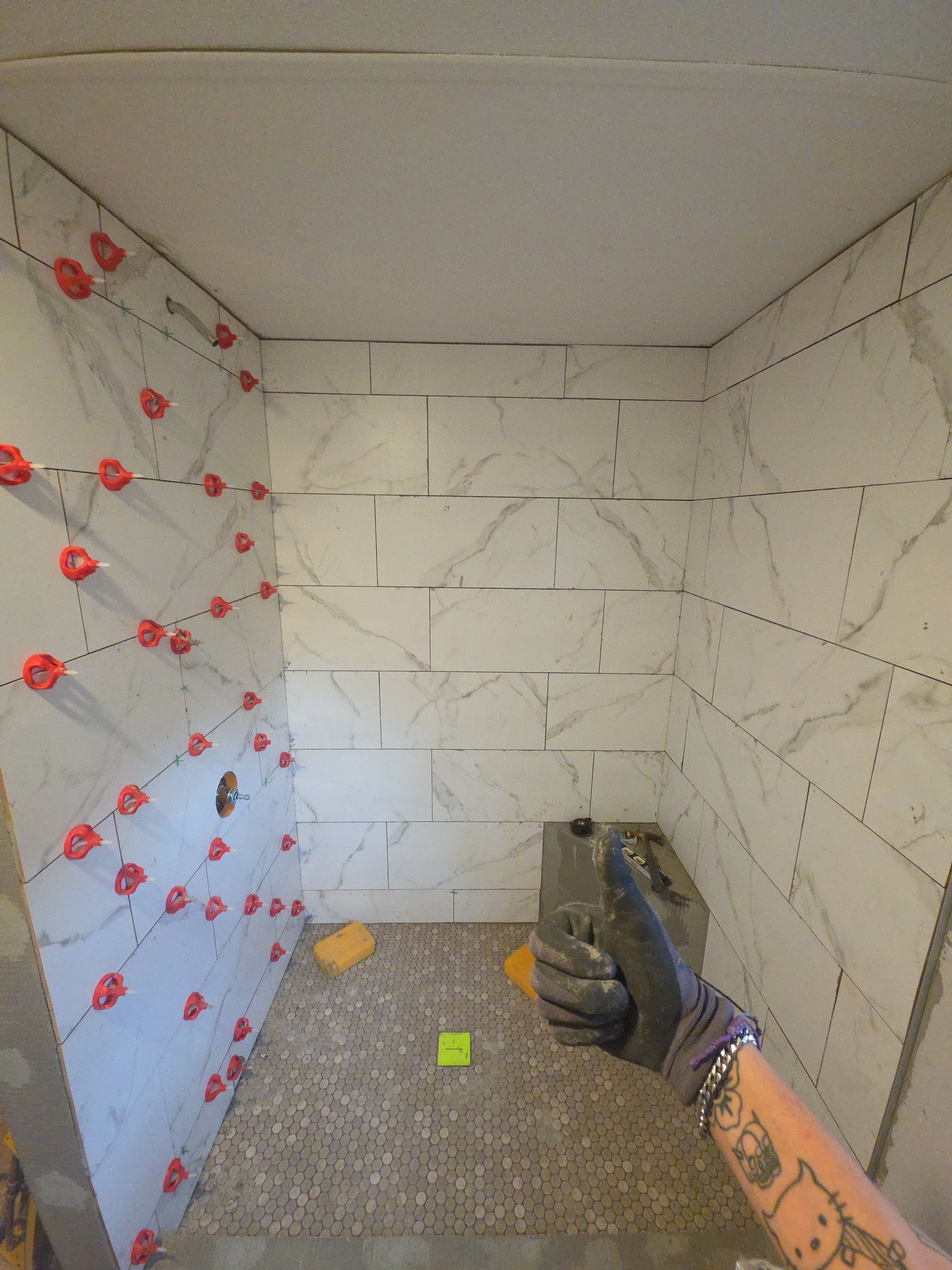 A person wearing gloves and a watch is holding a tile leveling system tool in a bathroom with ongoing tile installation. The bathroom has marble-patterned wall tiles and small circular floor tiles. There are spacers and leveling clips attached to the wall tiles.