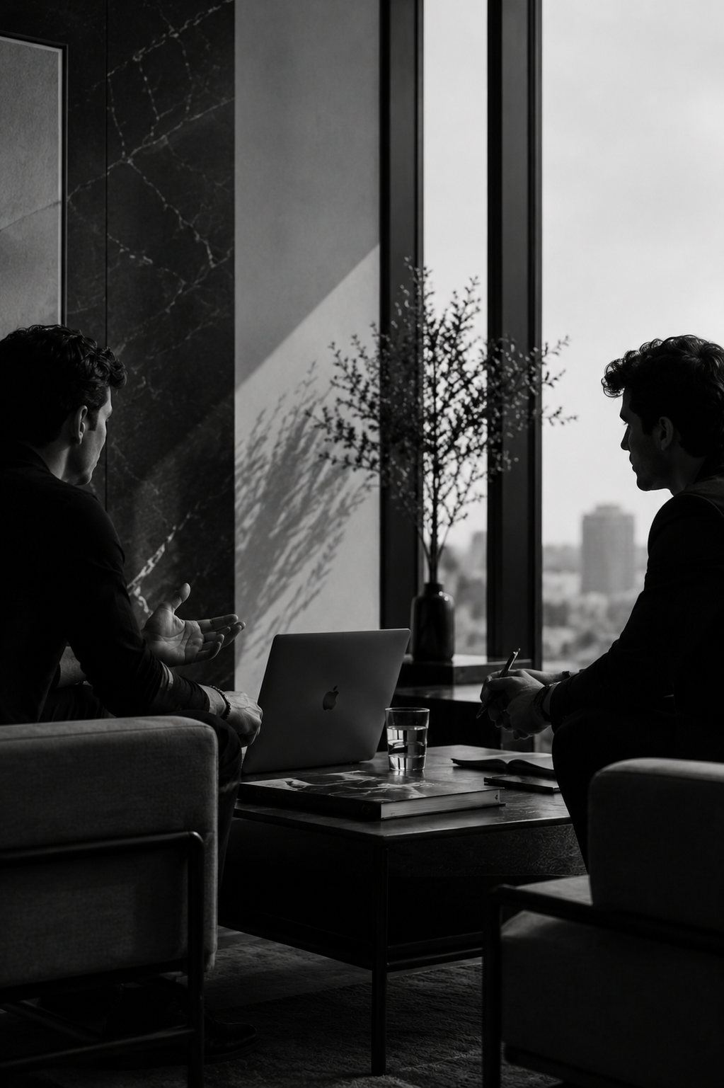 Two men having a serious conversation in an office lounge area, with a laptop, some books, and a glass of water on the table, near large windows showing a city skyline.
