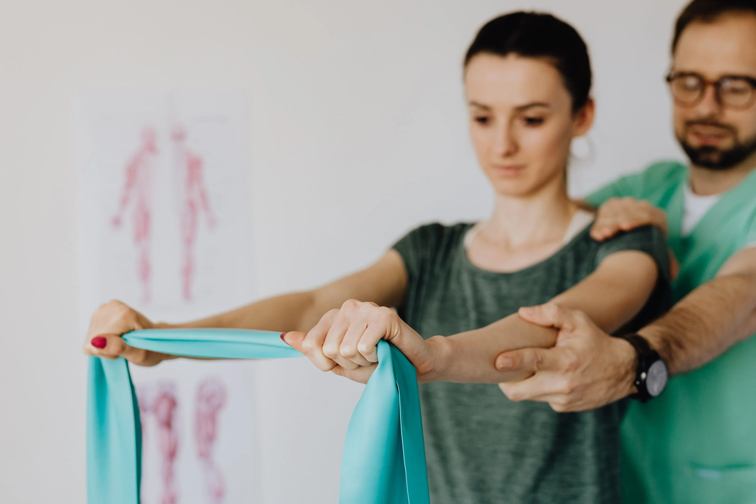 Two people, a woman with short dark hair and a man with glasses and a beard, are holding a blue resistance band during a physical therapy or exercise session. In the background, there is a poster of a human body silhouette with muscles marked.