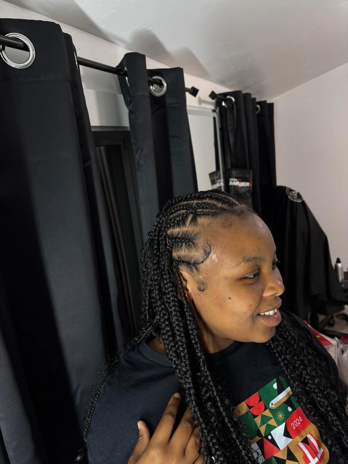 A woman with long box braids smiling while sitting beside black curtains, with a hand resting on her shoulder.