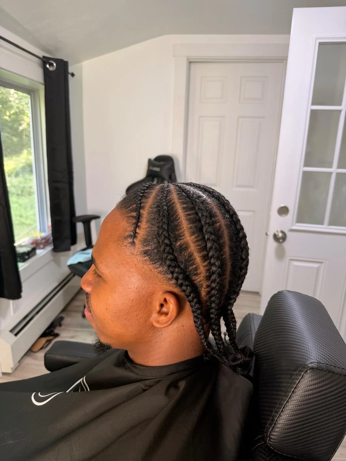 A man with braided hairstyle sitting in a barber chair in a room with white walls, a window with black curtains, and a white door. The room has natural light coming through the window.