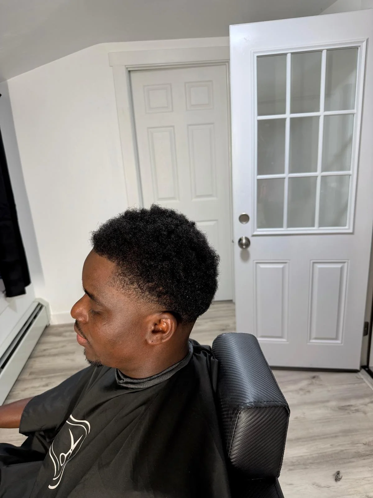 Profile view of a man with a fresh fade haircut sitting in a barber chair with a black cape, in a room with white walls, hardwood flooring, a white door with glass panels, and a closed closet door in the background.