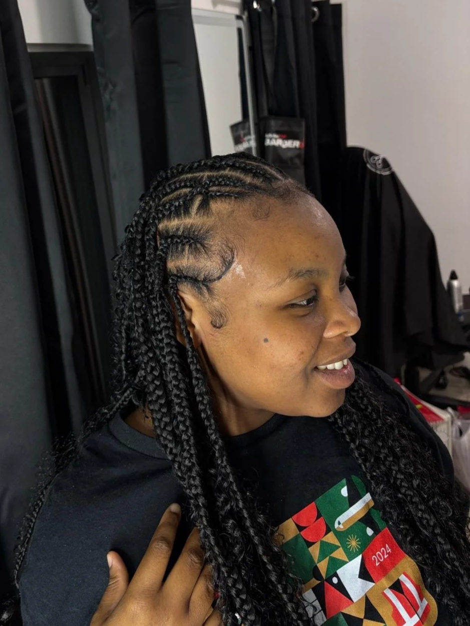 Woman with braided hair sitting in a salon chair, smiling, with hair styling tools and salon equipment visible in the background.