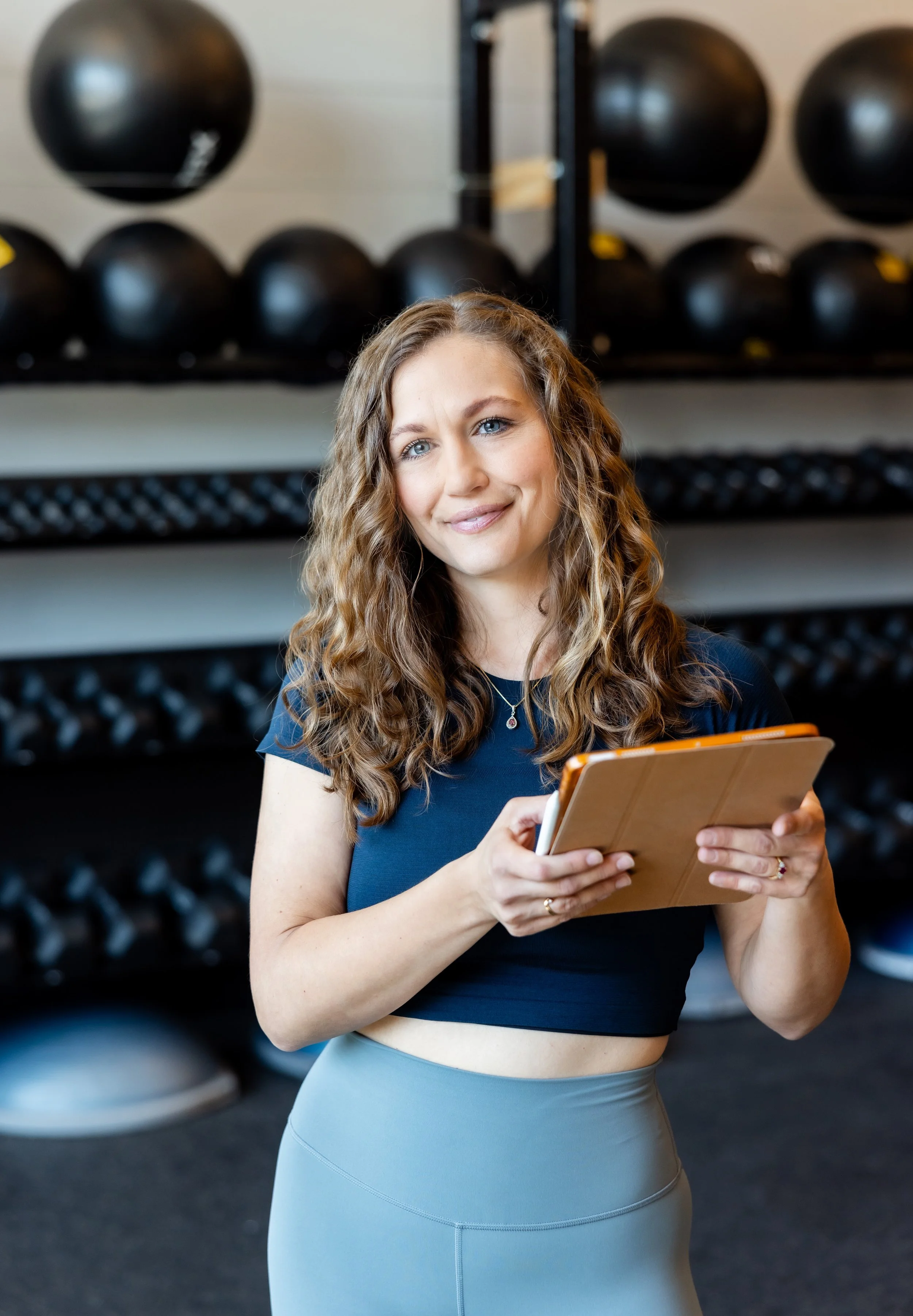 A woman holding a clipboard in a gym with black medicine balls on shelves in the background.