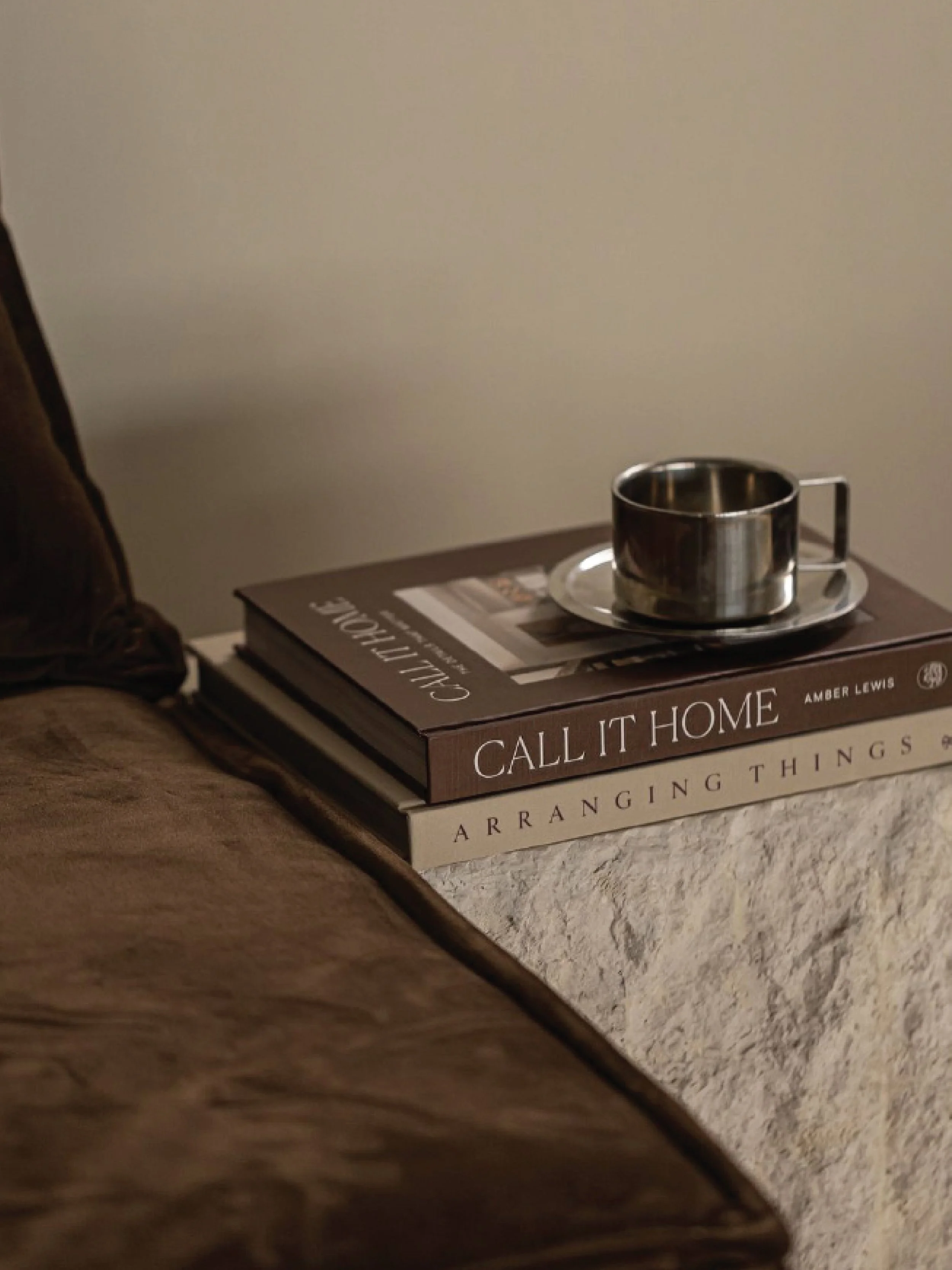 A stack of three books on a wooden surface with a metal cup and saucer on top, positioned next to a beige wall.