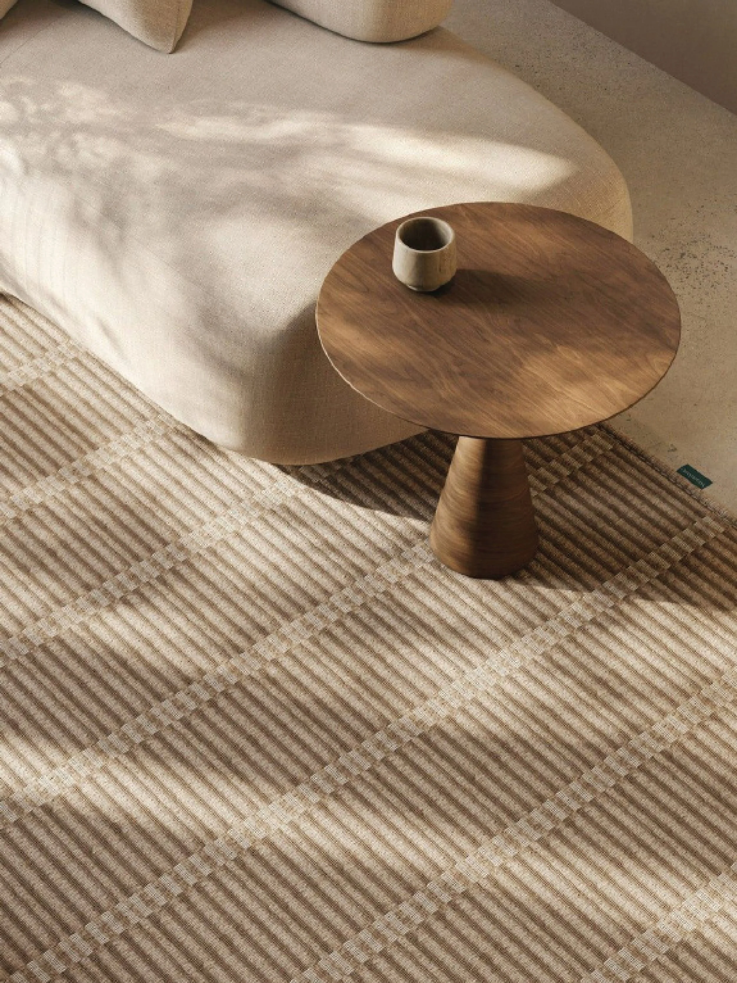 Close-up of a wooden table with a ceramic cup on it, placed on a beige striped carpet next to a beige sofa.