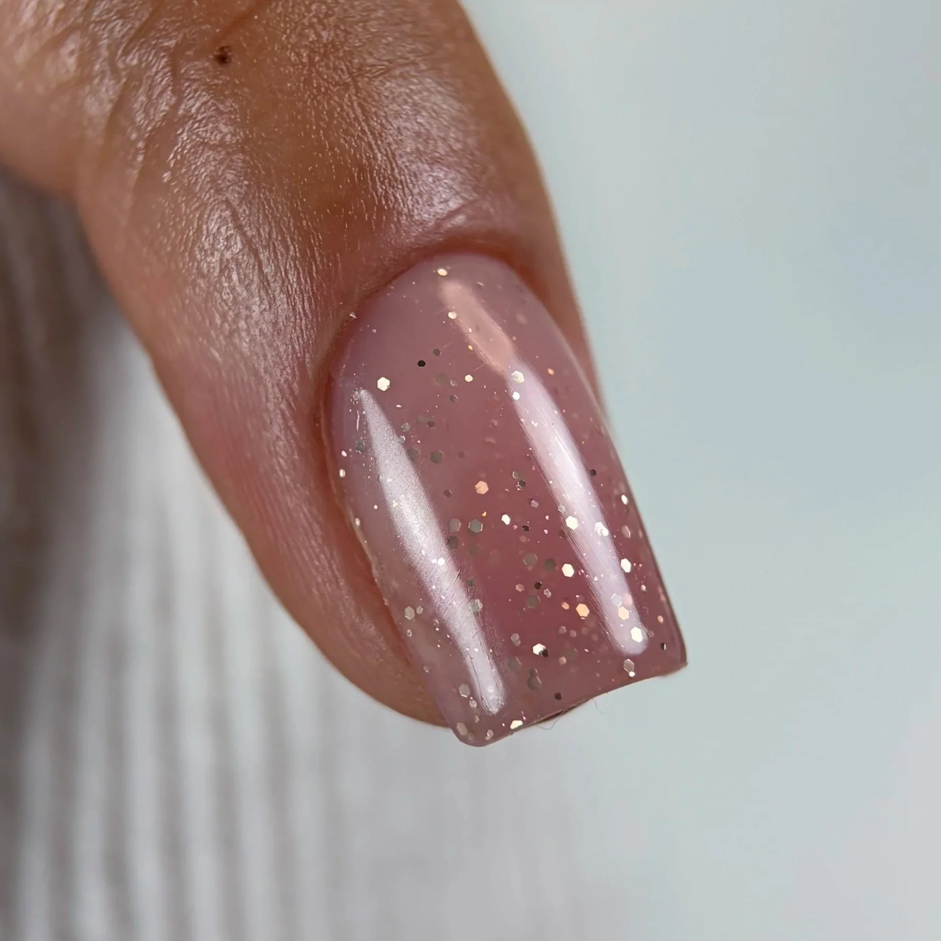 Close-up of a person's finger with pink nail polish that has gold glitter, and a section of skin with some fine skin texture and small dark spots.