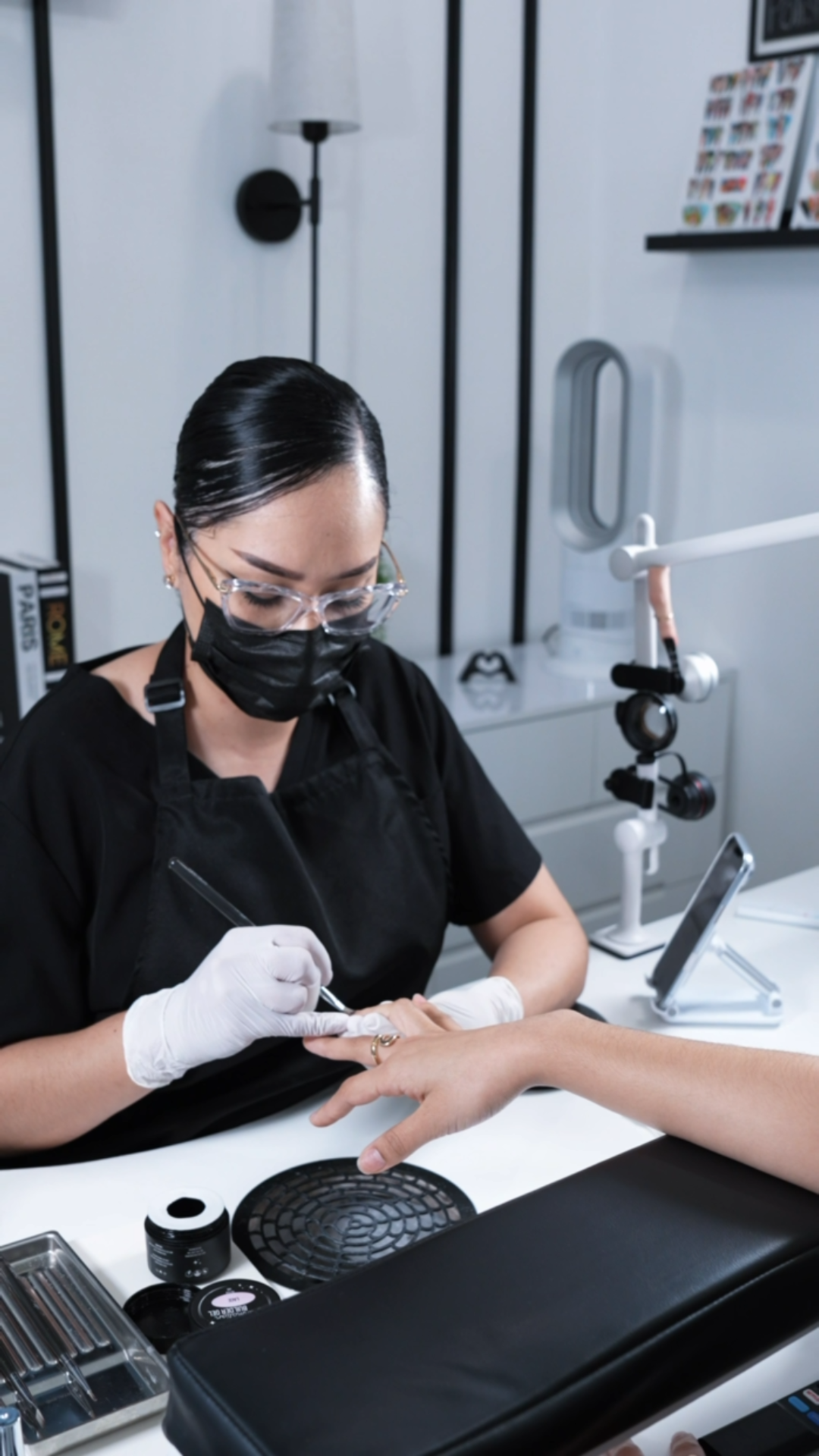 A woman wearing protective glasses, mask, and gloves doing a manicure or nail art at a professional salon station with tools and a digital device nearby.
