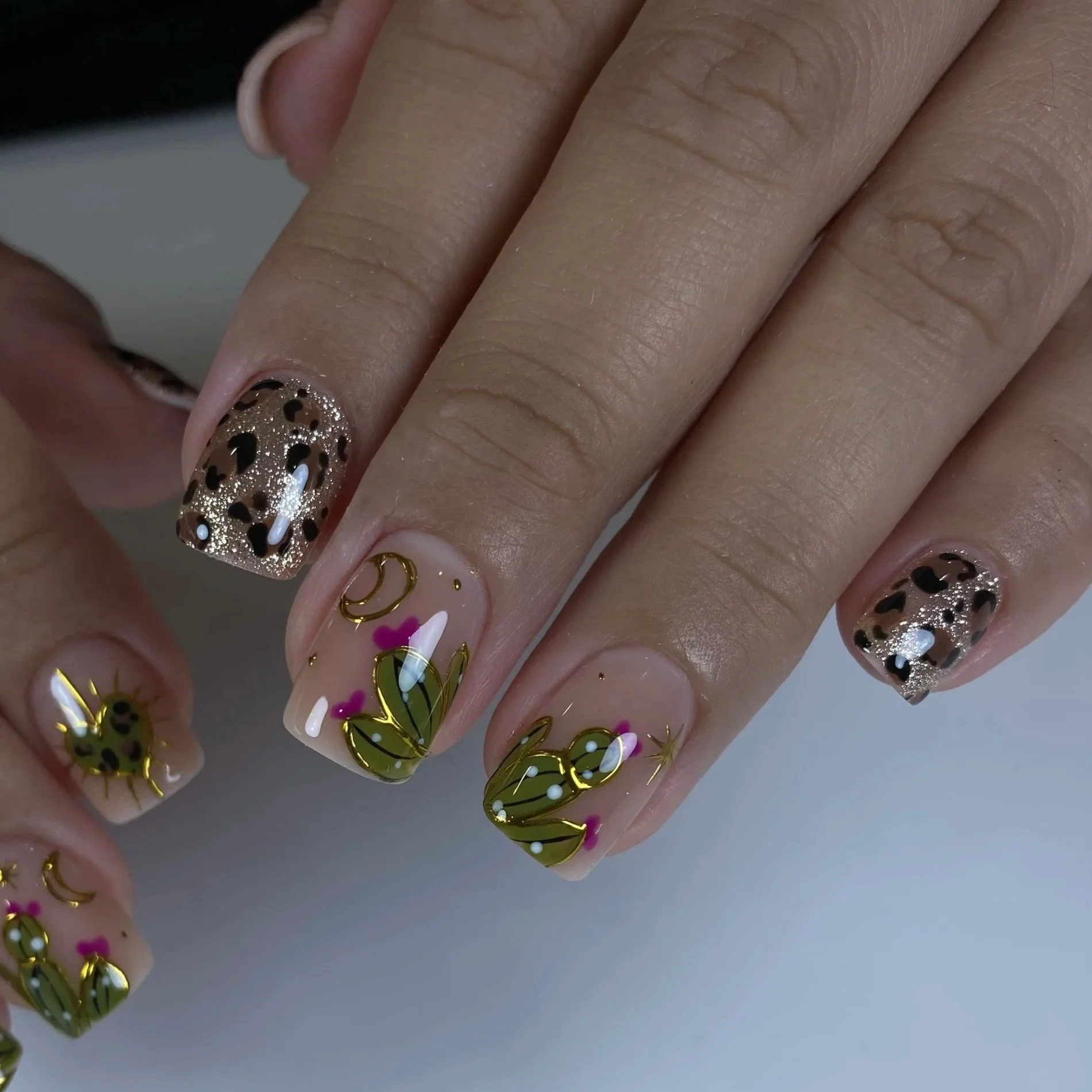 Close-up of a hand with decorated fingernails featuring gold and black leopard print, floral patterns, and accents of pink and white, with visible skin texture and knuckles.