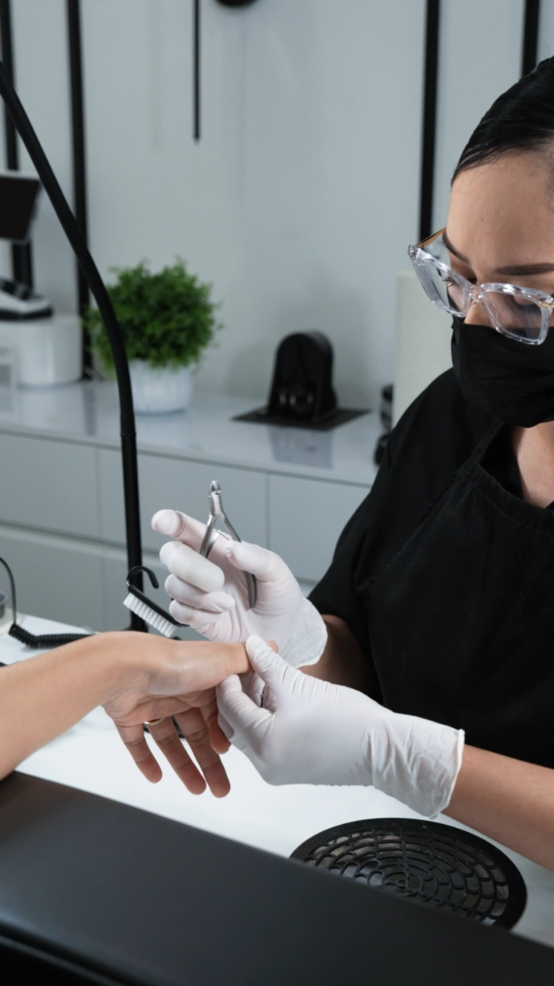 A woman in protective glasses, a face mask, and gloves is doing  a manicure with gloved hands, in a nail studio setting with a white counter and a potted plant in the background.