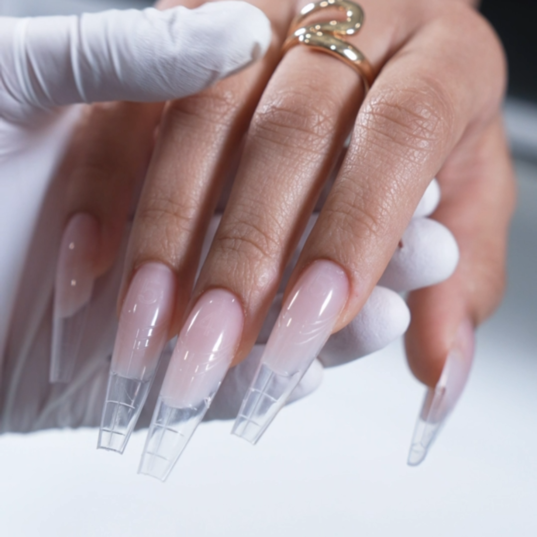 Close-up of a hand with long, square-shaped acrylic nails being manicured, wearing gold rings, with a gloved hand holding the fingers